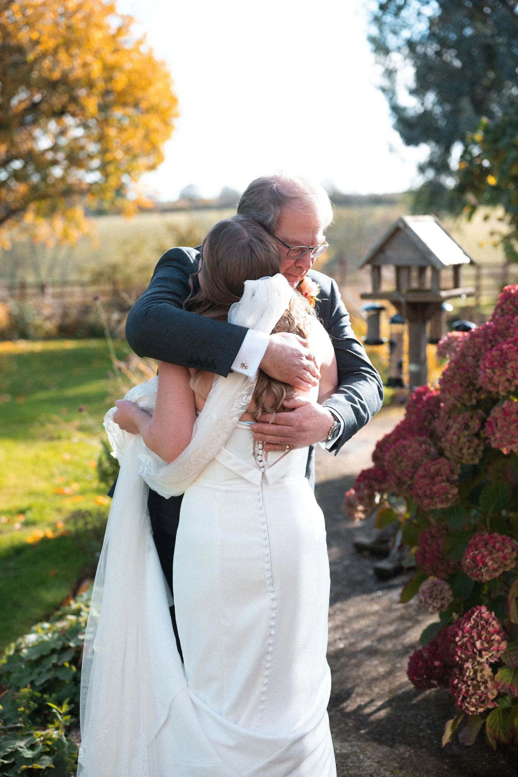 A wedding scene with a bride and an older man, likely her father, hugging outdoors during fall. The bride is in a white wedding dress with a long veil, and the scene features colorful fall foliage and hydrangea bushes.