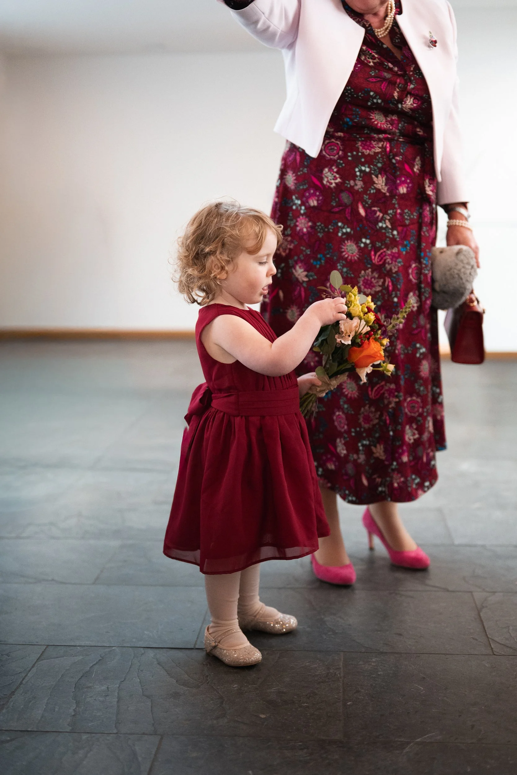 A young girl with curly blonde hair wearing a red dress, beige tights, and glittery shoes, holding a bouquet of flowers at a formal event.
