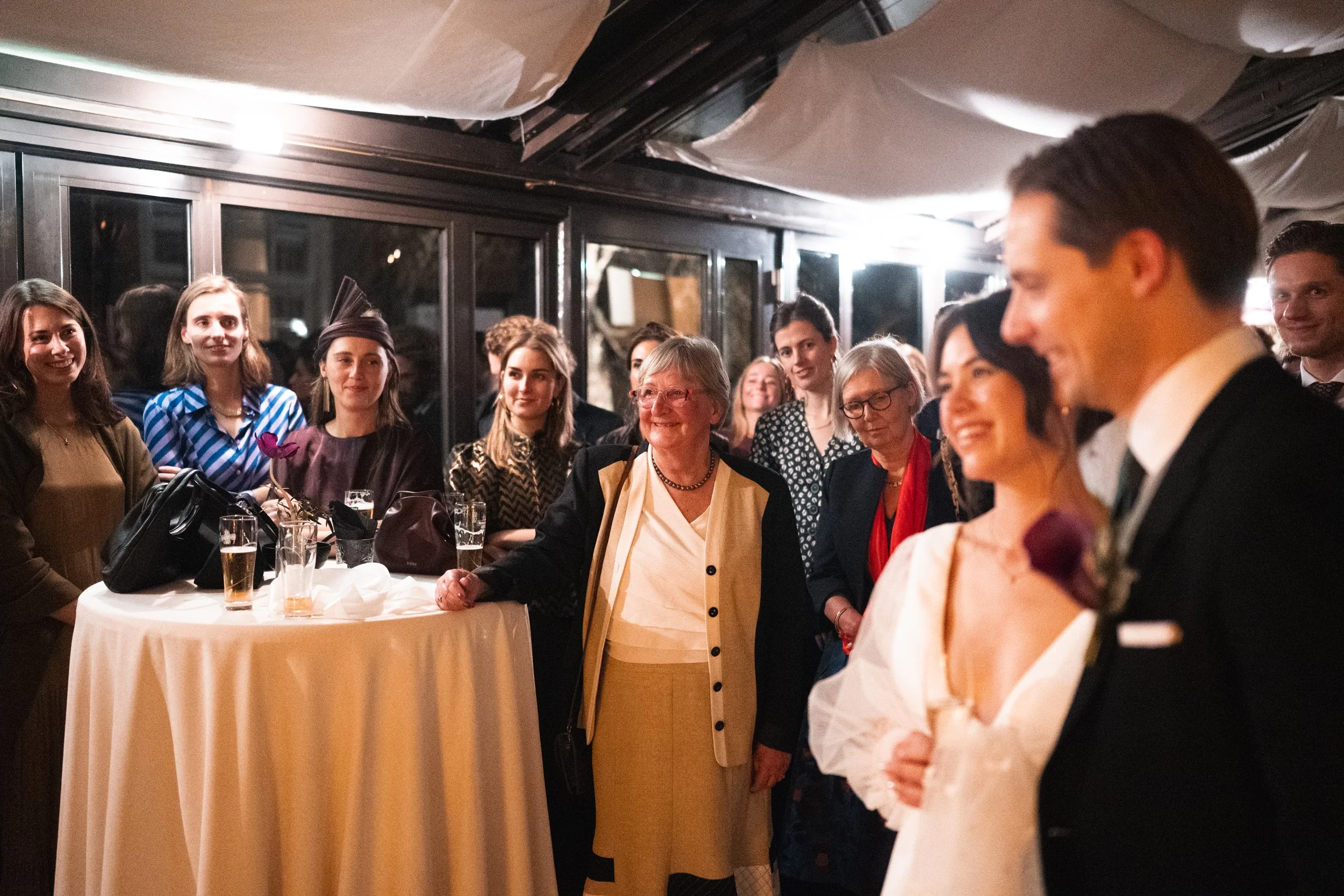 People gathered at a wedding reception, with the bride and groom in the foreground and guests smiling around a high-top table in the background.