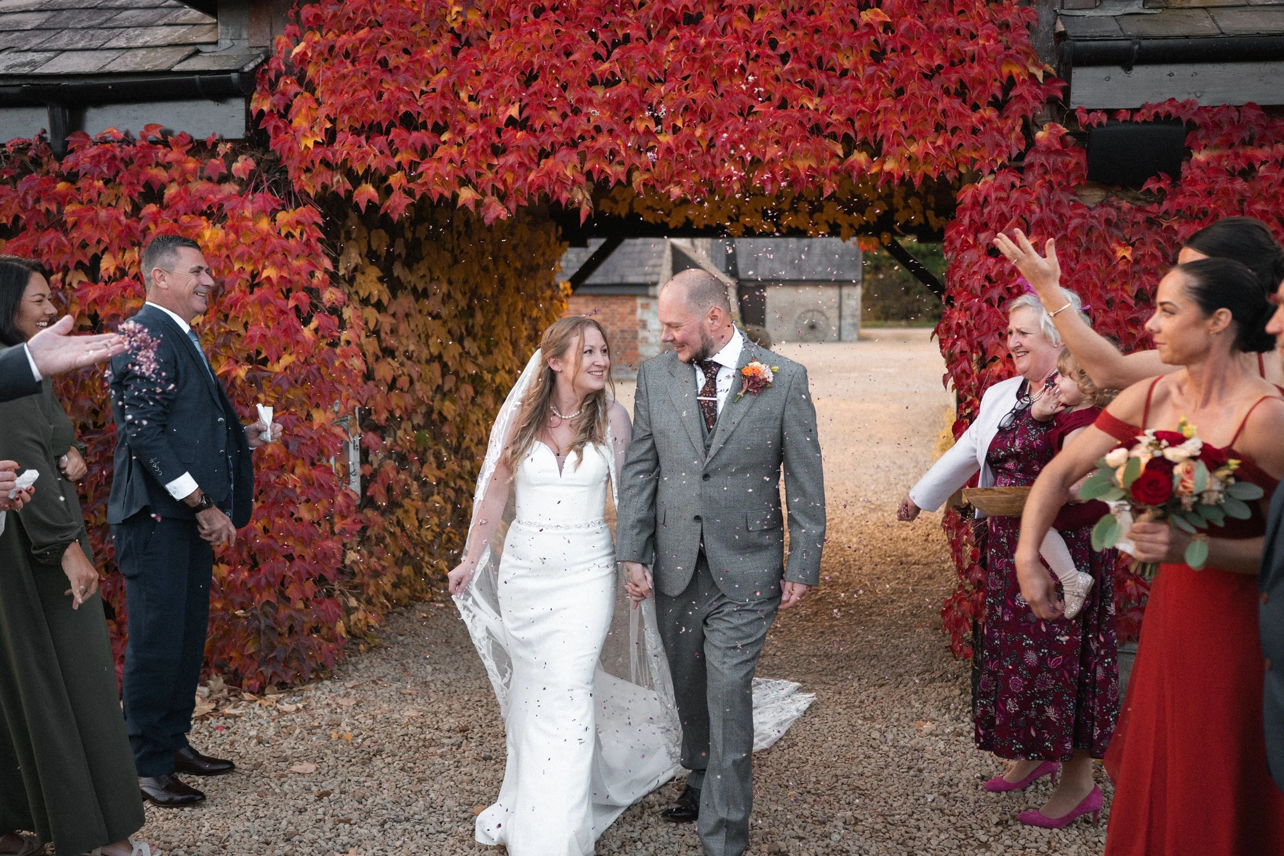 A newlywed couple walks hand in hand through a confetti shower surrounded by friends and family outdoors under red autumn foliage.