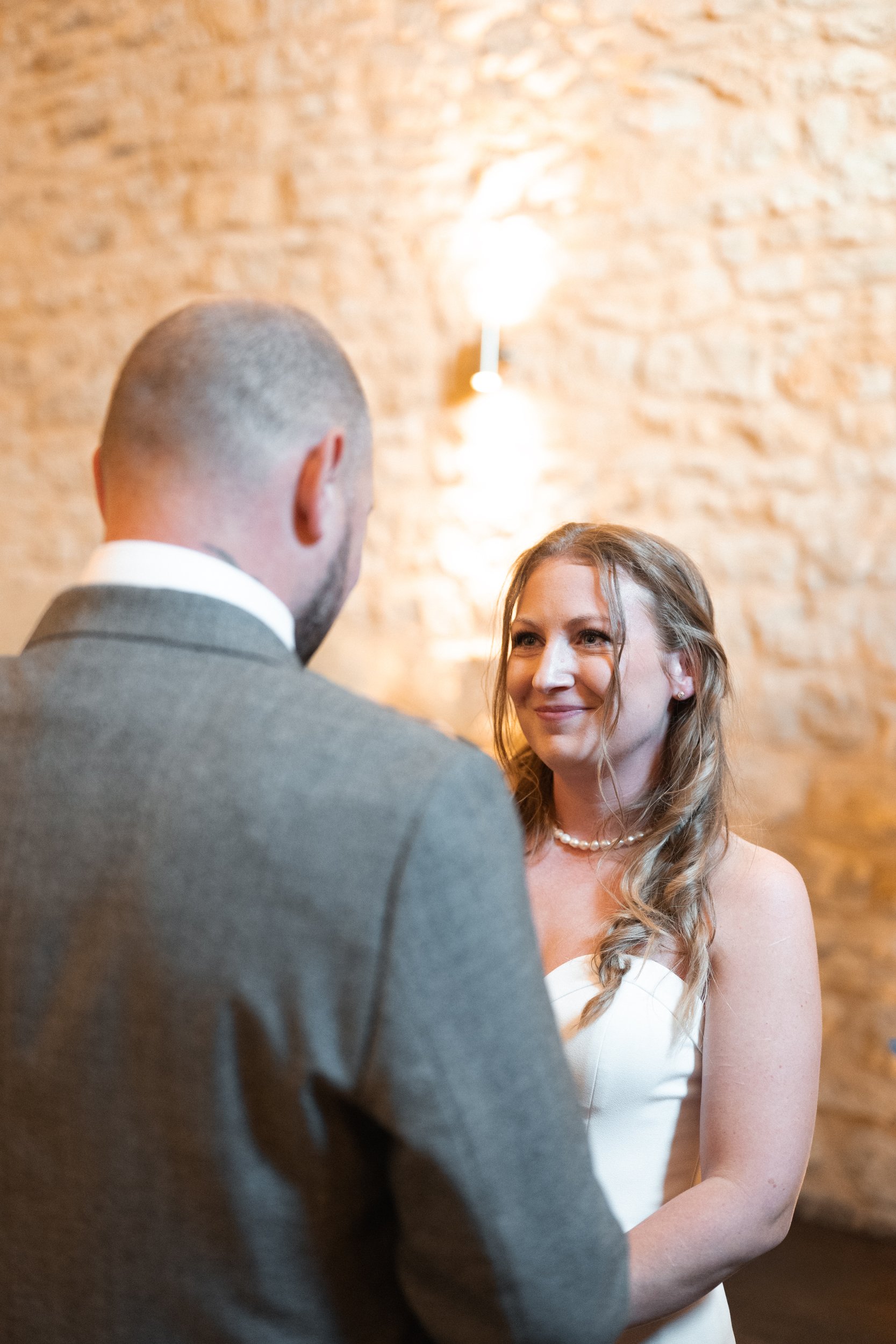 A bride and groom exchange vows indoors. The bride is smiling at her partner, wearing a strapless white wedding dress, with wavy hair and a pearl necklace. The groom, seen from the back, wears a gray suit and has a closely shaved haircut. The backgro