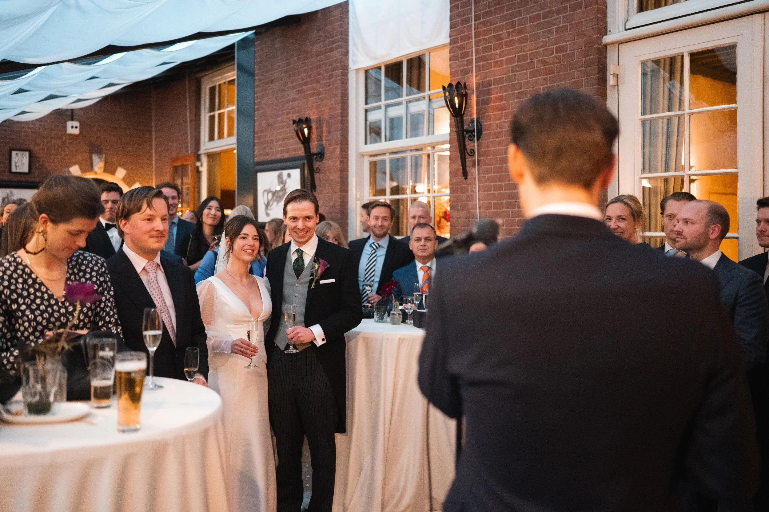 Wedding reception with guests listening to a speech, including the bride and groom smiling, indoor venue with brick walls, large windows, and warm lighting.