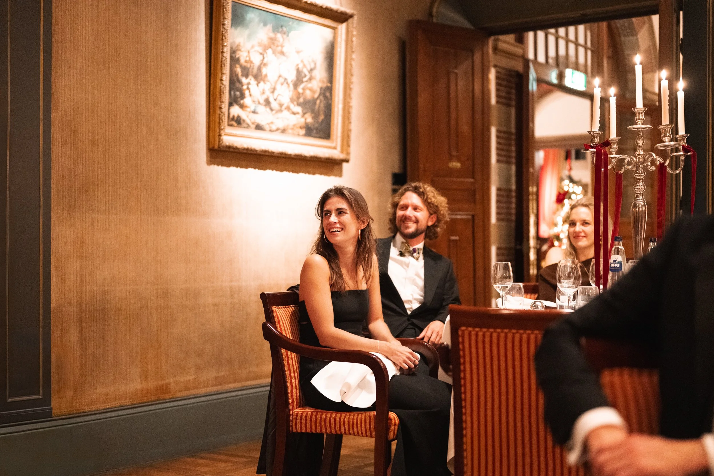 A group of people enjoying a formal dinner at a restaurant, with a woman and man smiling at a table with wine glasses and a candelabra in the foreground.