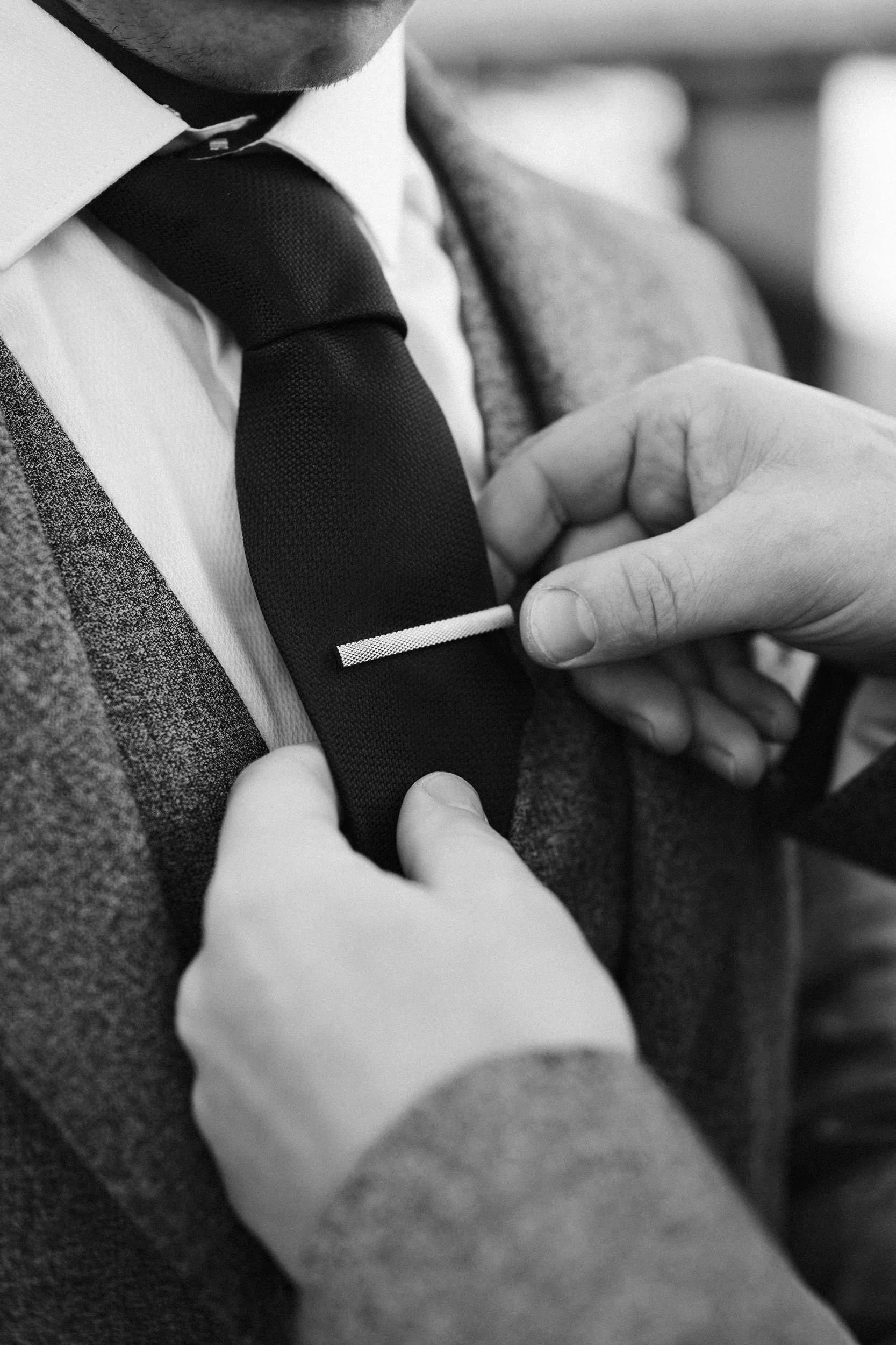 Person adjusting a tie on a man dressed in a suit, white shirt, and plaid blazer, black tie, in black and white.