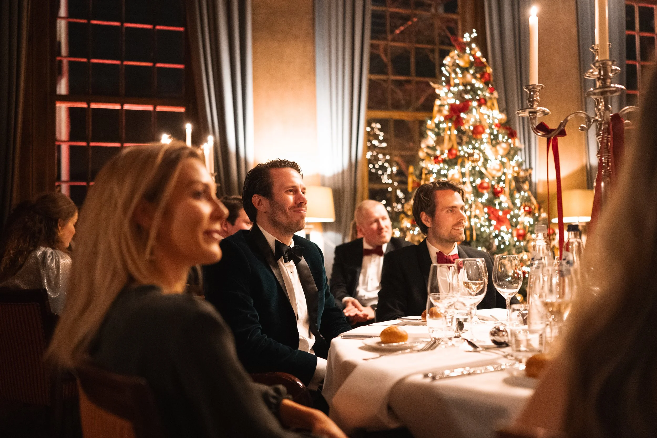 People in formal attire sitting at a Christmas dinner table with a decorated Christmas tree in the background, indoors at night.