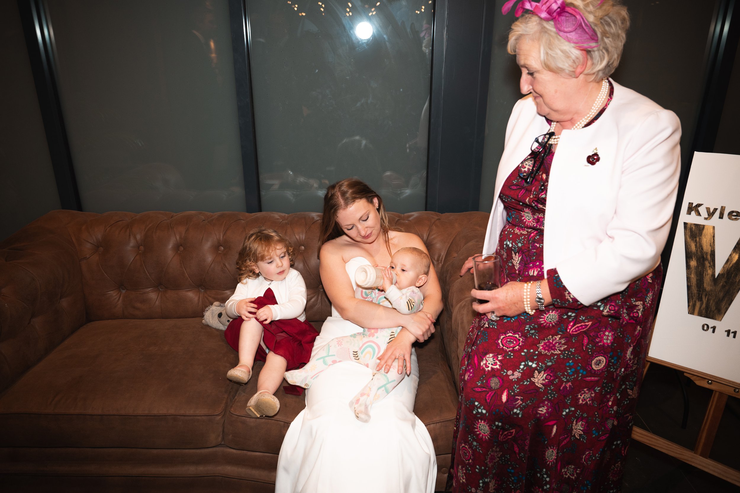 Elderly woman with pink headband and white blazer standing next to a young woman with long brown hair sitting on a brown leather couch, breastfeeding a baby. A toddler girl with curly hair and a red dress is sitting on the couch, holding a stuffed an