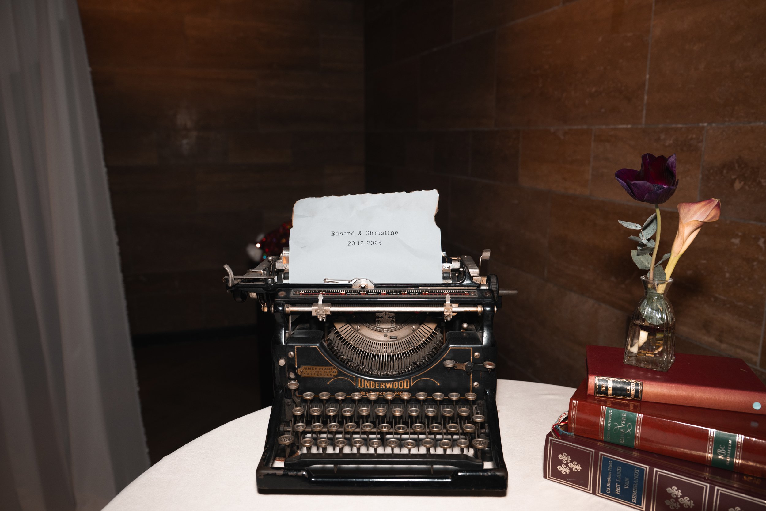 Vintage typewriter with a note on it, next to a small vase with purple and peach flowers, and a stack of books on a white table.