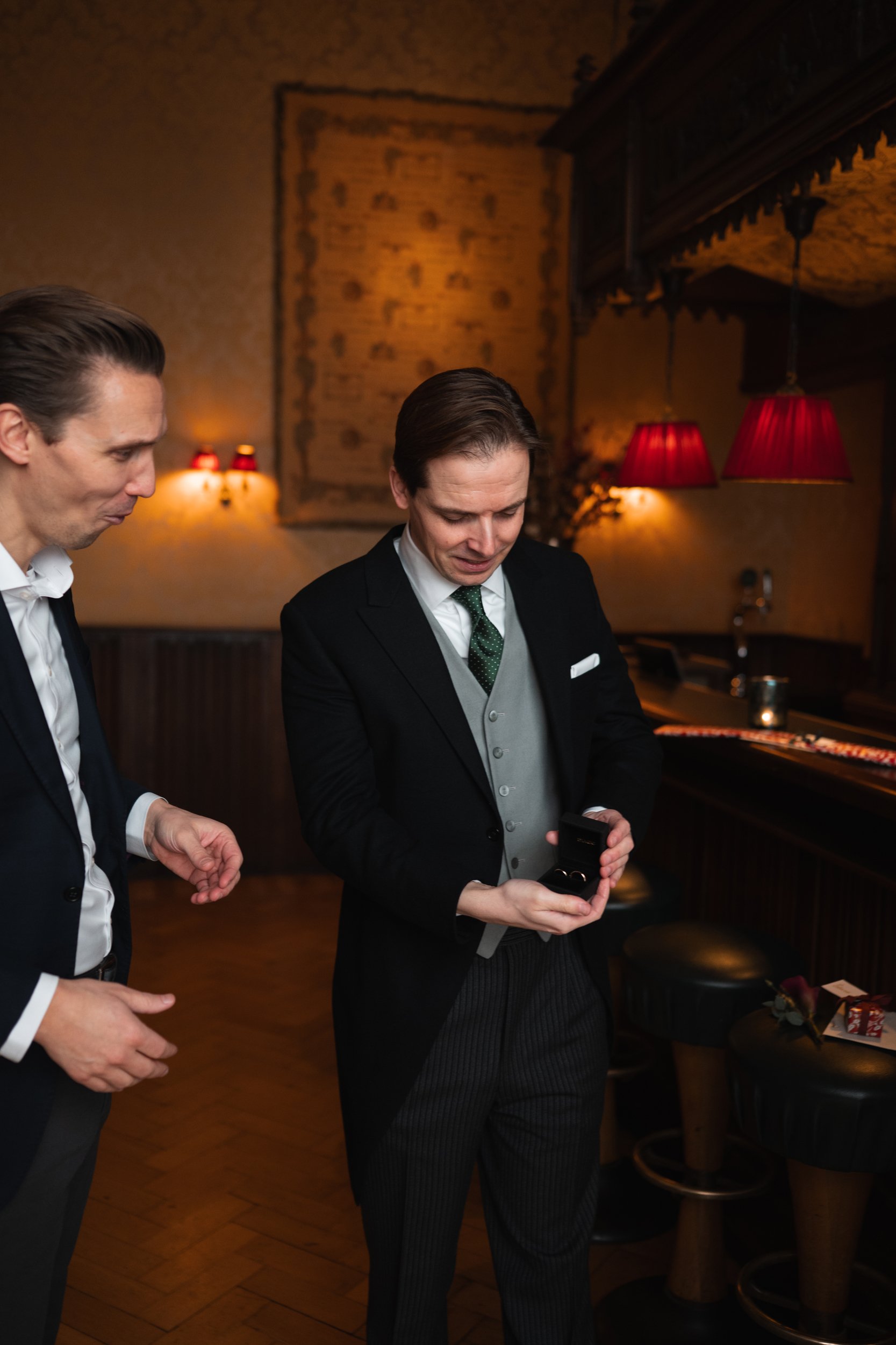 Two men in formal suits are standing in a dimly lit restaurant. One man is holding a small black box, likely an engagement ring box, and the other man is looking at it. The background features warm lighting, a bar counter, and red lampshades hanging 