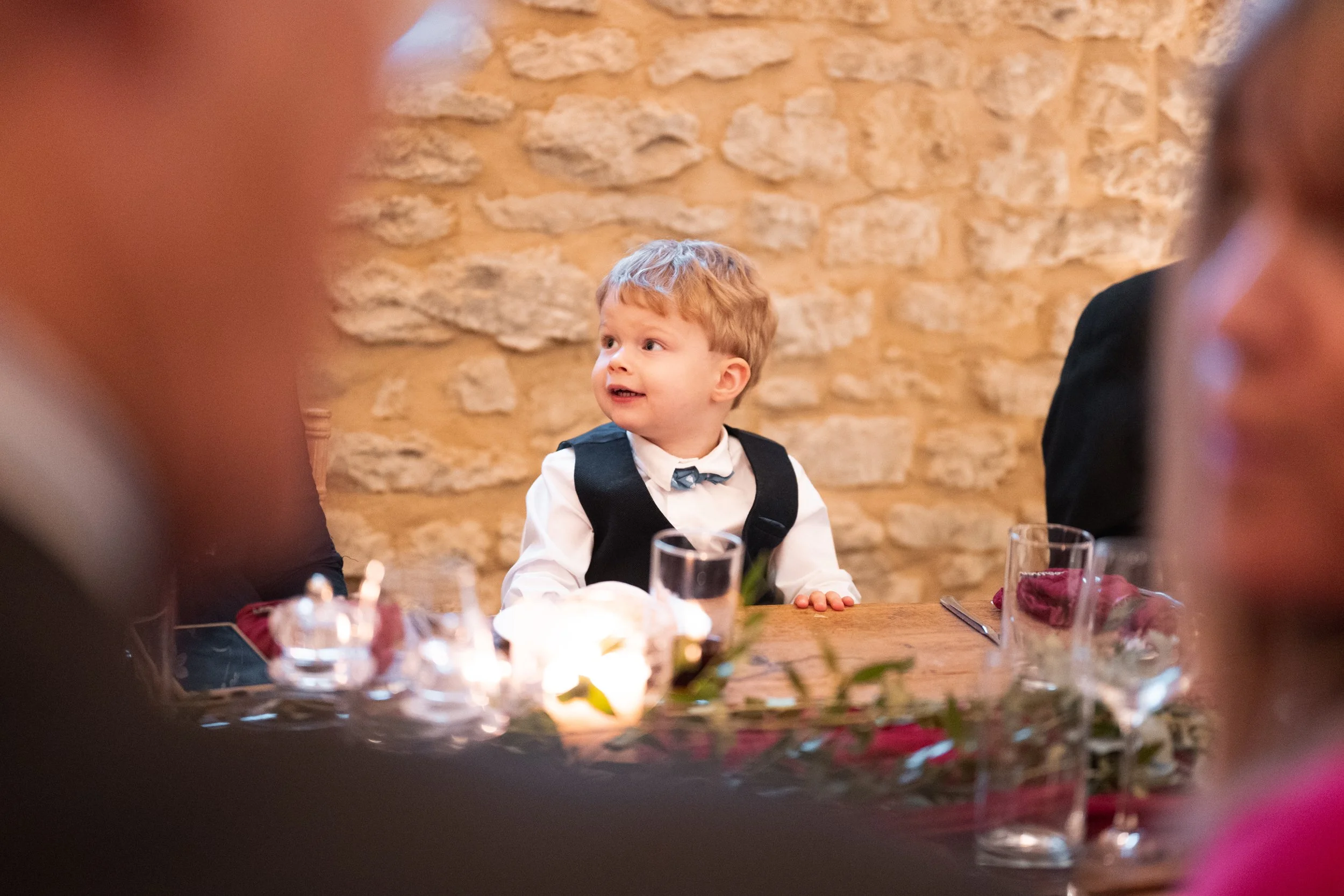 A young boy dressed in formal attire, including a white shirt, bow tie, and vest, sitting at a dinner table in a rustic setting with a brick wall background, surrounded by other people at the table.