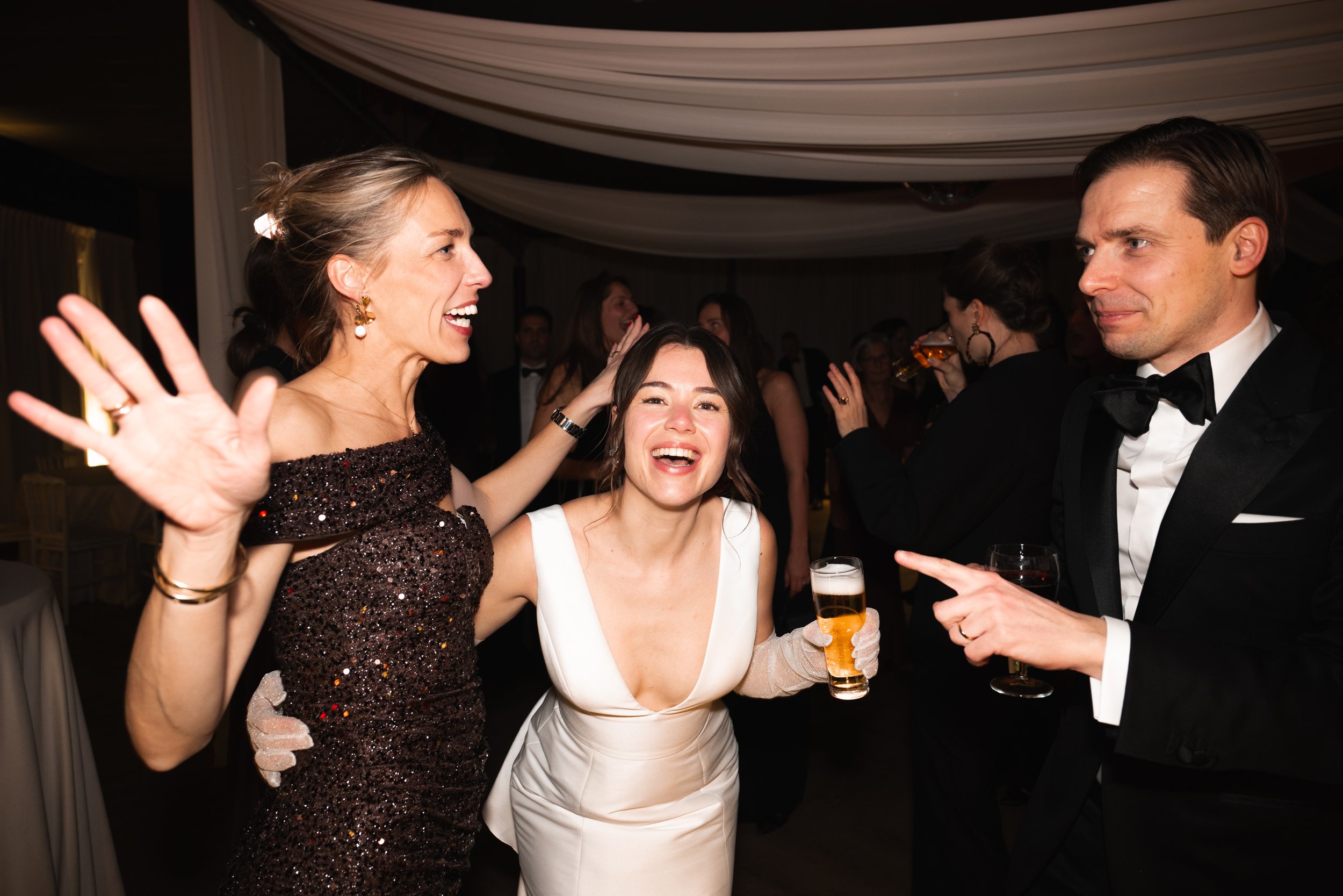 Three people cheer and converse at an elegant event, with women in dresses and a man in a tuxedo, surrounded by others socializing under a decorated ceiling.