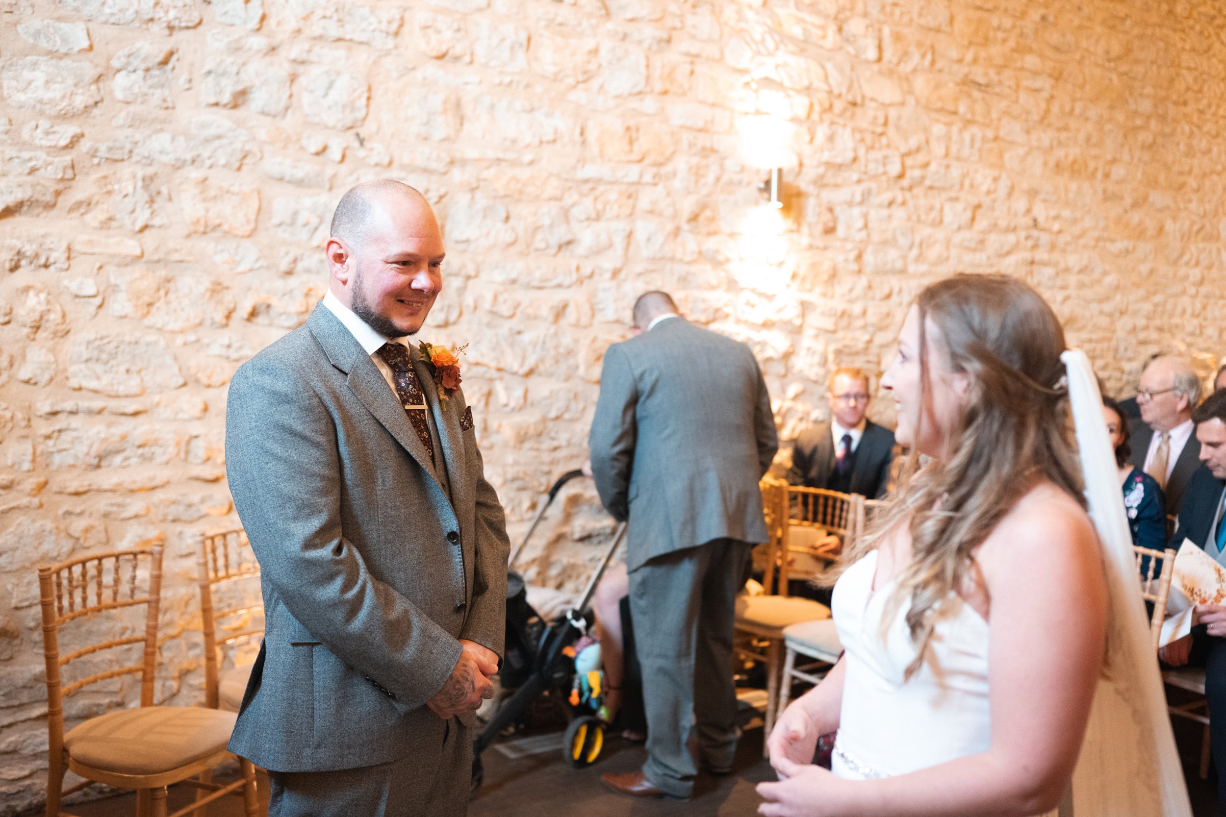 A man and woman are smiling at each other during a wedding ceremony in a rustic stone-walled venue. The man is dressed in a gray suit with a boutonniere, holding his hands together. The woman is in a white dress with a veil, looking at the man. Guest