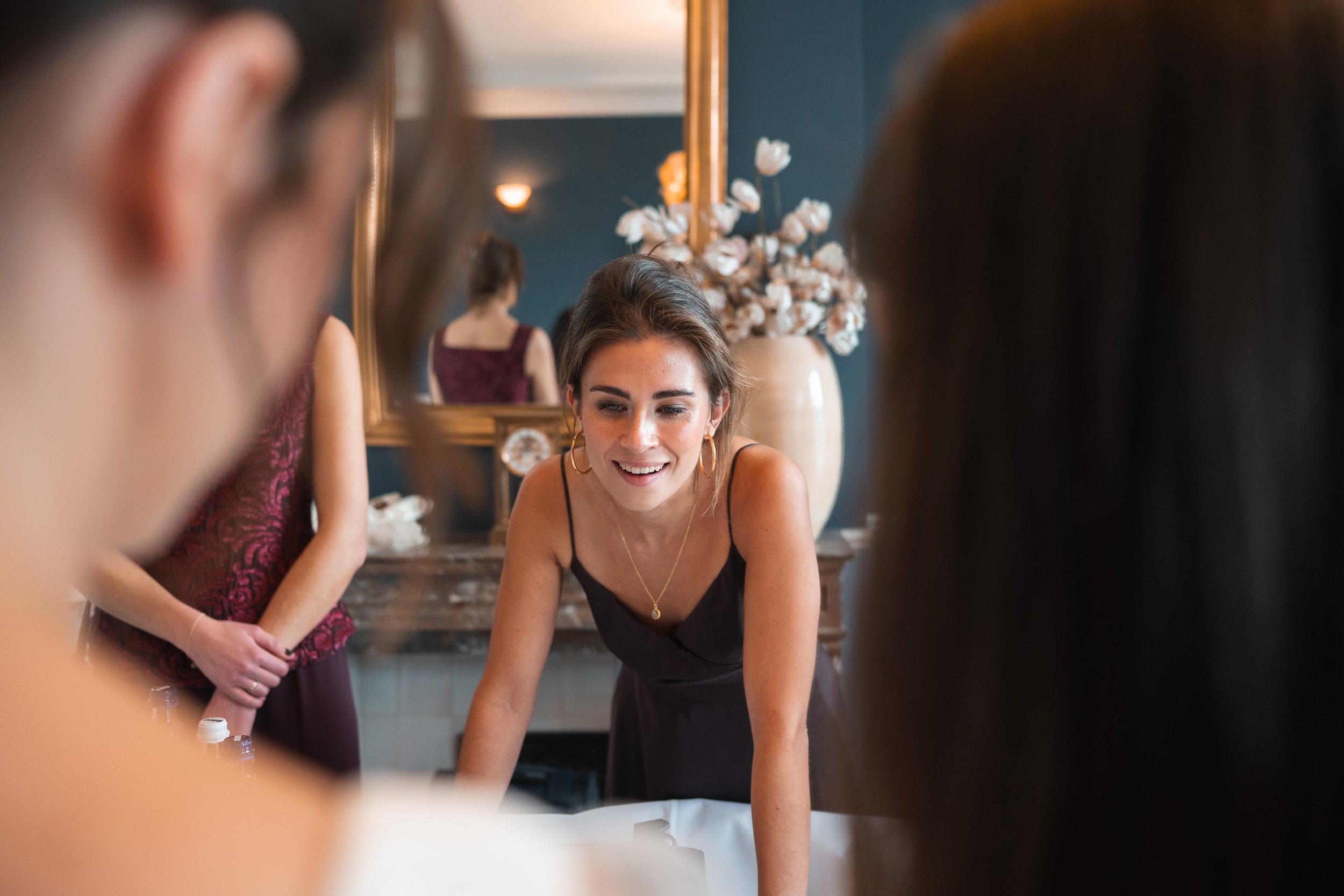 Woman smiling and leaning forward, surrounded by others, in a room with a large vase of white flowers and a mirror in the background.