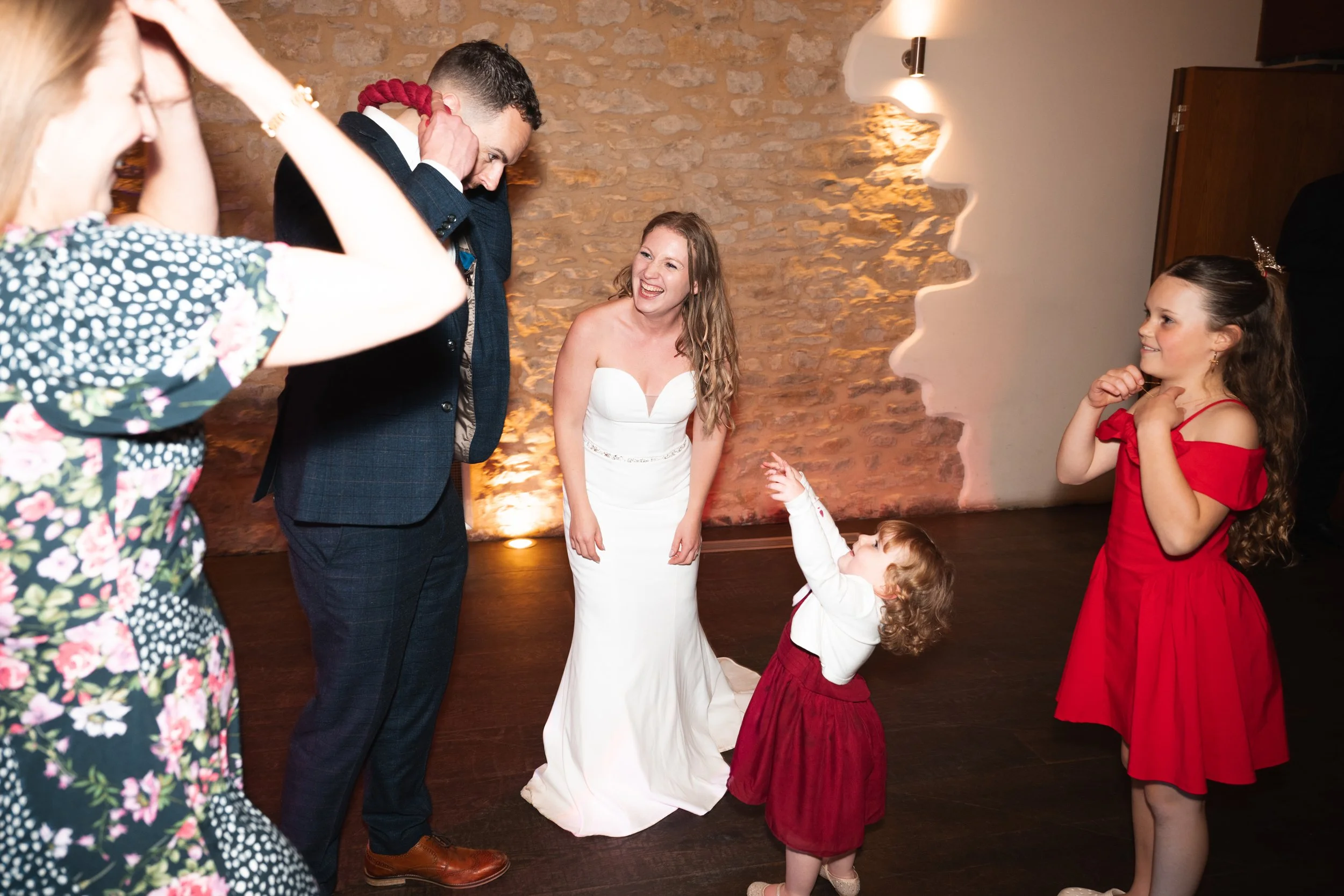 A bride in a white wedding dress laughing while standing on a dance floor with children and adults. A young girl in a red dress is reaching for a man wearing a dark suit, holding a red rope. Two other girls, one in a red dress and one in a floral dre