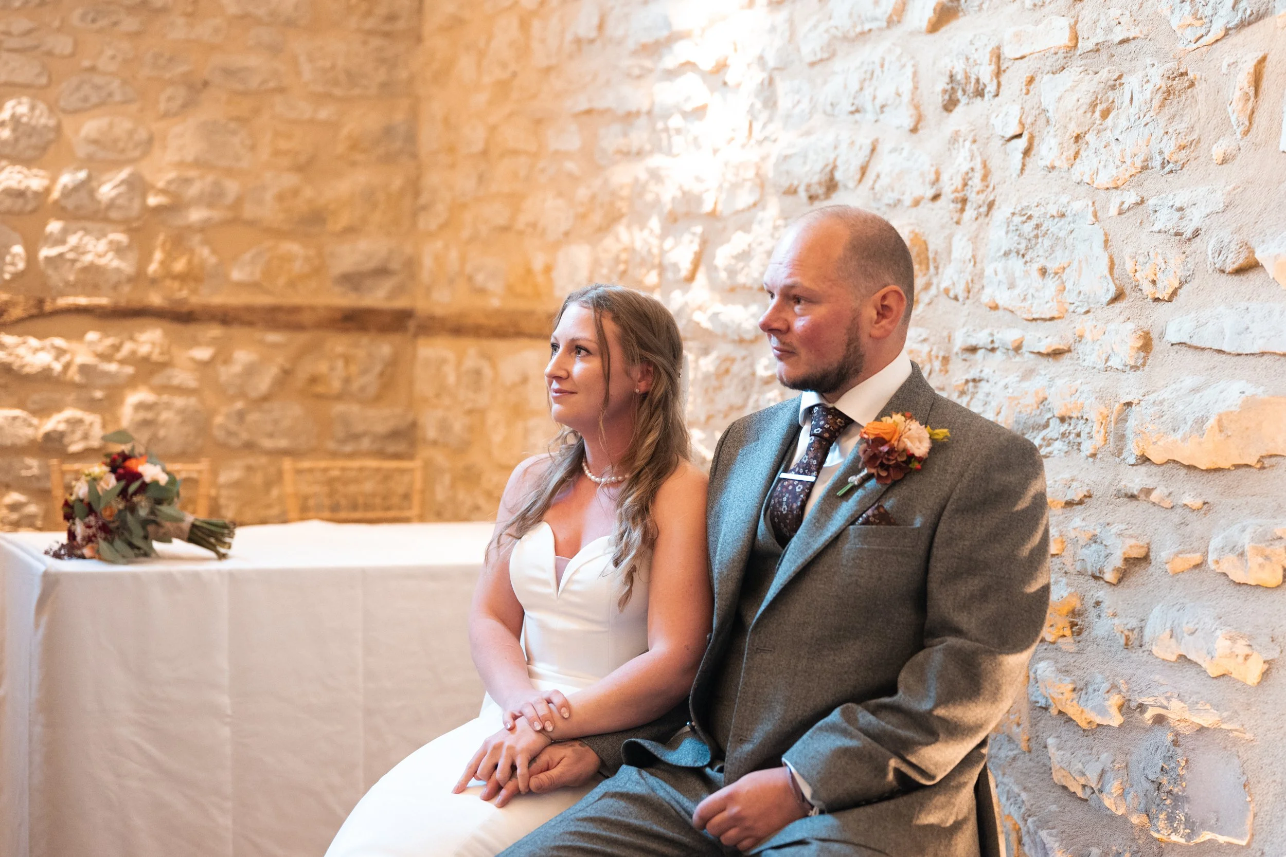 A bride and groom sit side by side during their wedding ceremony inside a stone-walled room, appearing attentive and content.