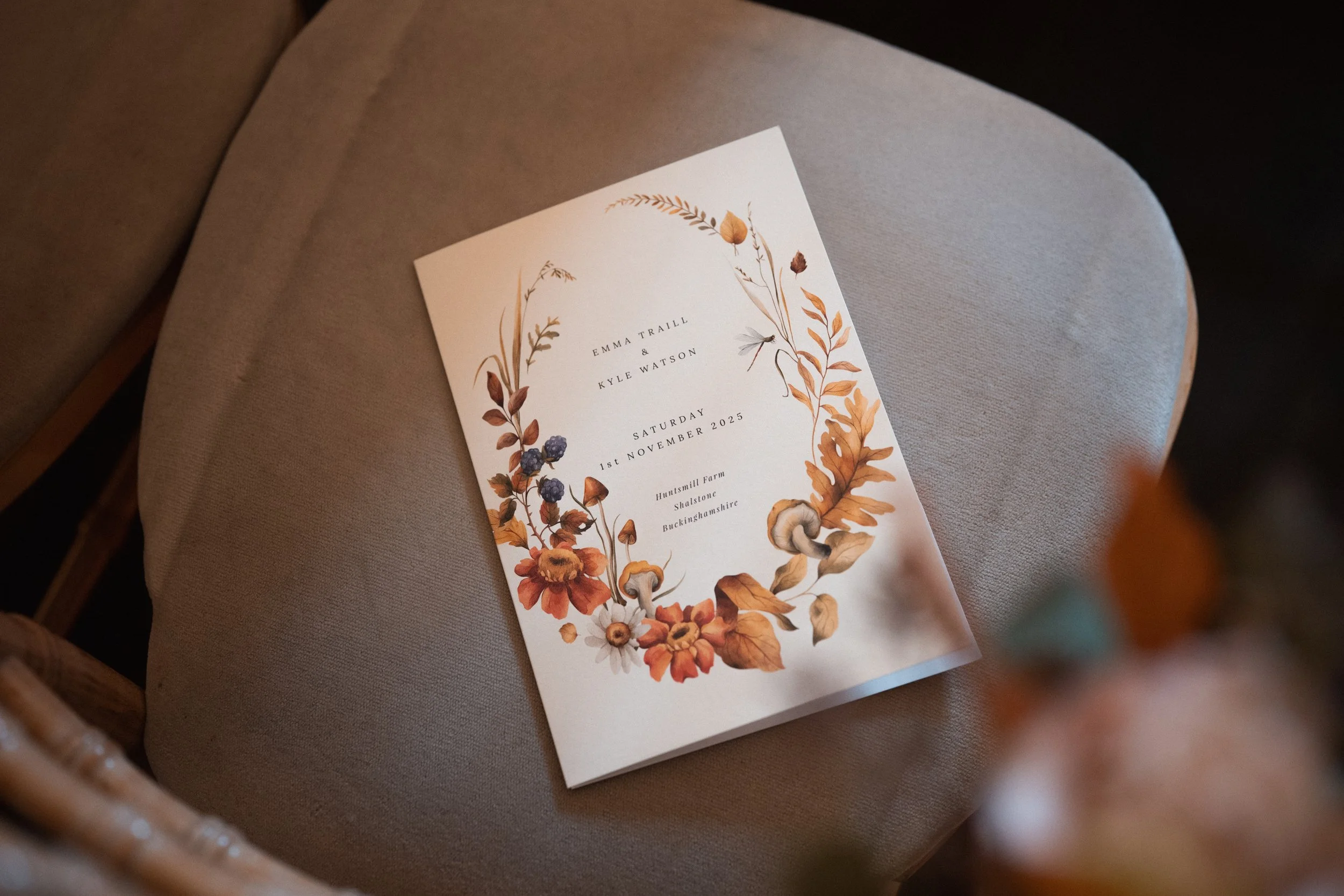 Wedding invitation with floral design, featuring orange, brown, and dark purple flowers, and text with details of the ceremony on a white background.