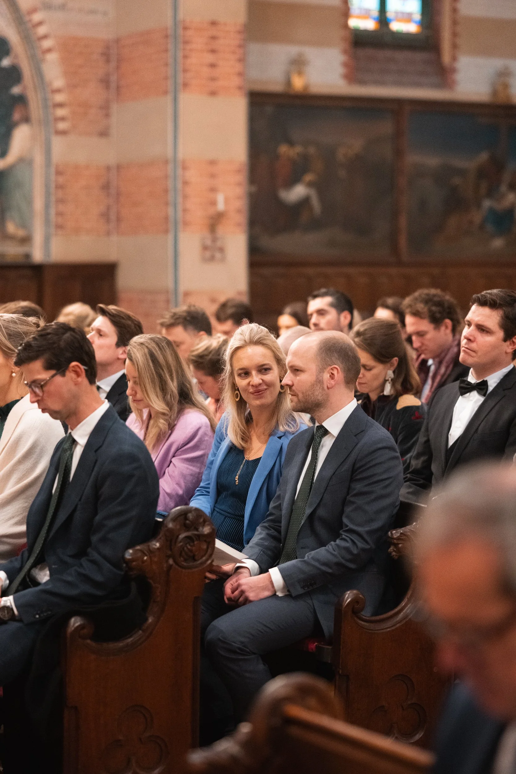 People sitting in wooden pews inside a church, dressed in formal attire, attending a ceremony or service.