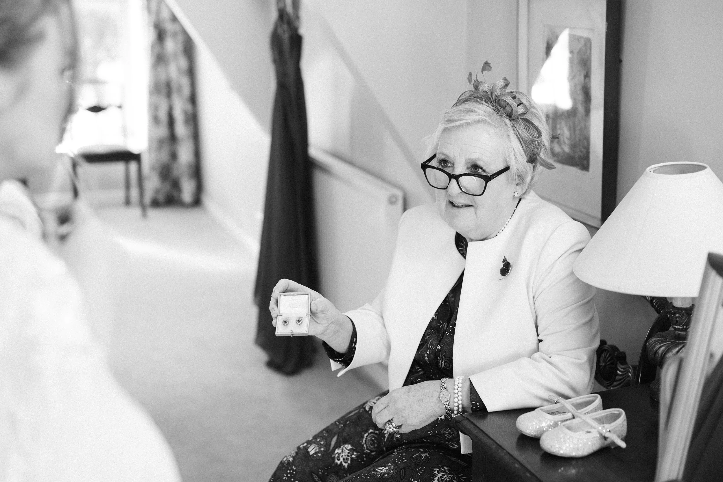 An elderly woman with glasses and a floral headband shows a pair of earrings in a jewelry box to a person on her left in a room decorated with curtains, artwork, a table lamp, and a pair of glittery shoes.