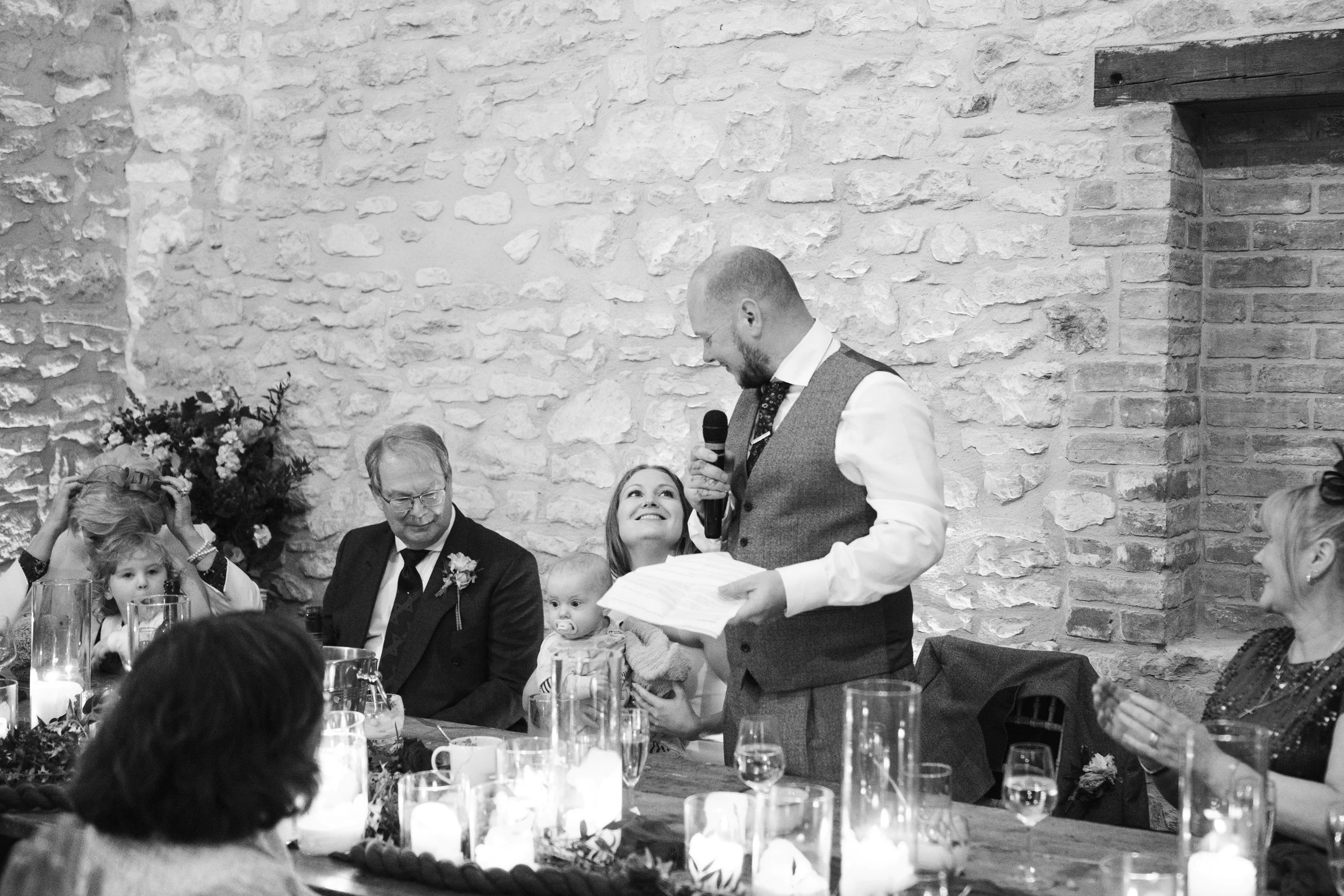 A man giving a speech at a wedding reception, holding a microphone and paper, while a woman with a baby looks up at him. Guests at a decorated table with glasses and candles are watching, sitting against a white brick wall.