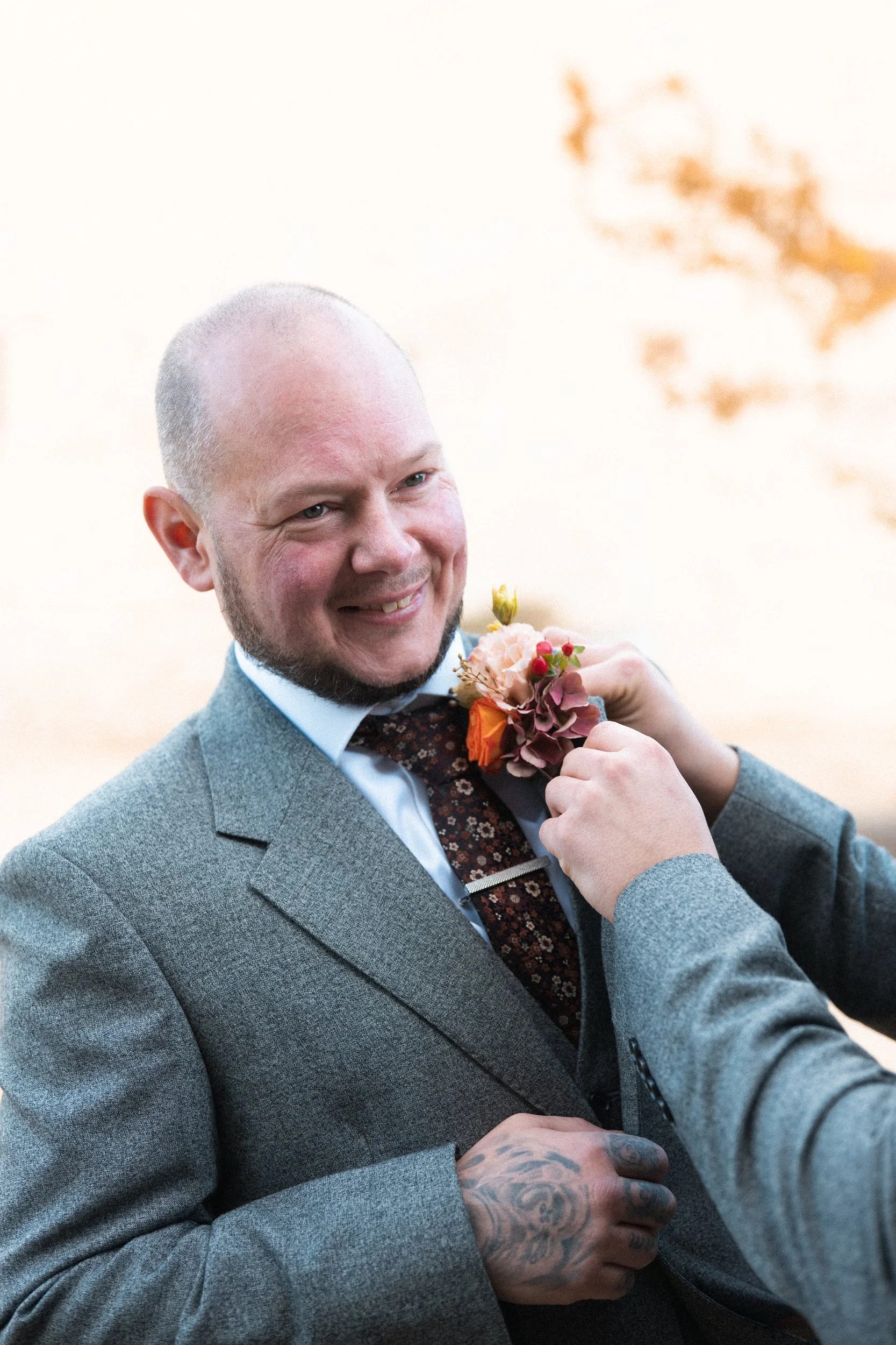 A man in a gray suit with a floral tie is smiling as another person adjusts a flower boutonniere on his lapel. The background is blurred with soft warm tones.