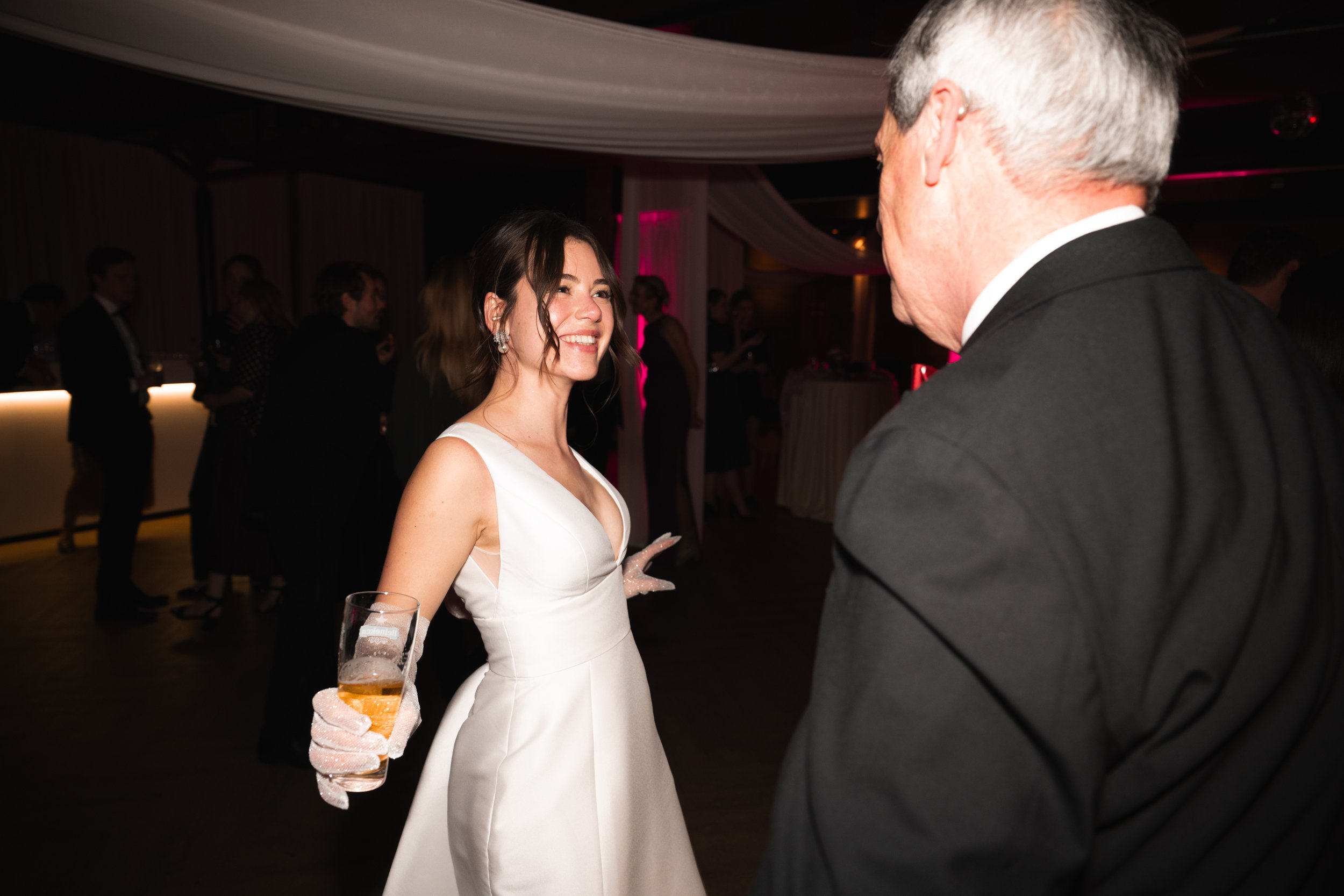 A woman in a white dress holding a drink, smiling, and talking to a man in a black suit at a party or wedding reception.