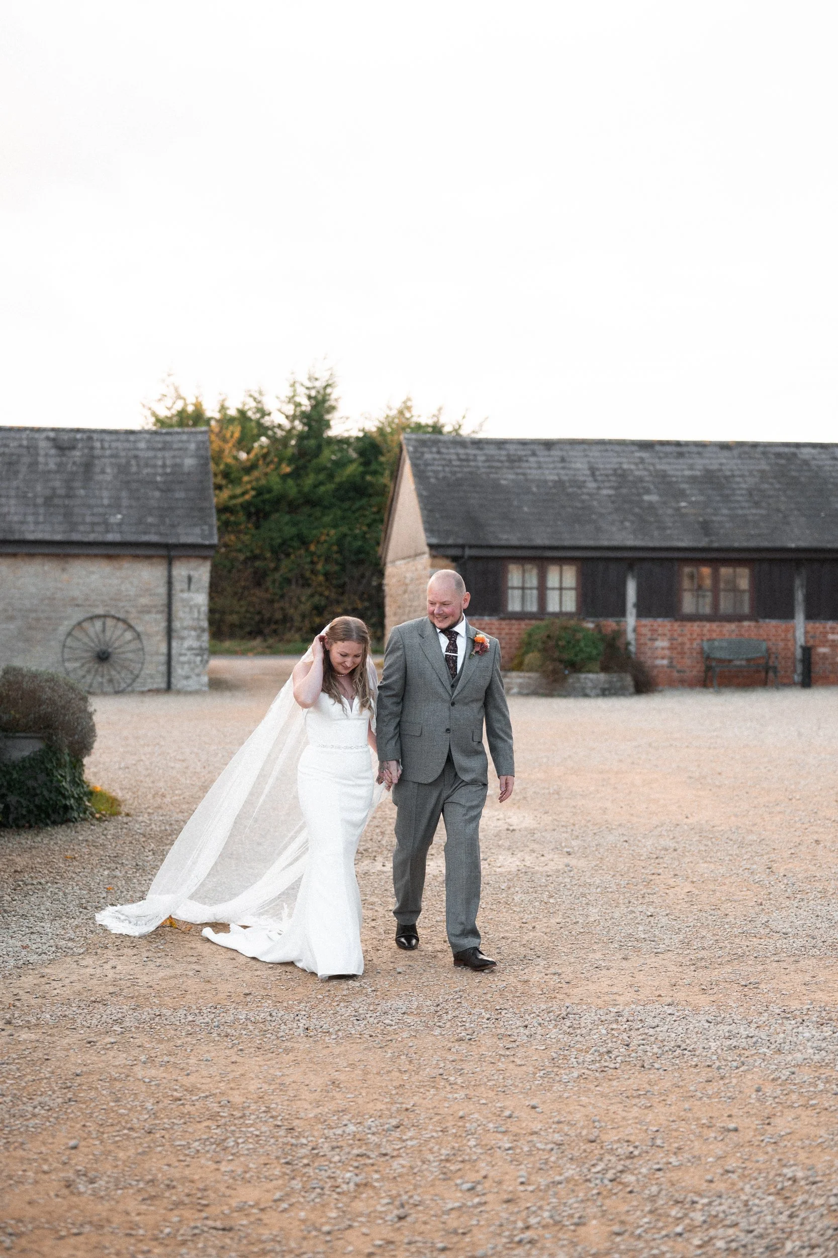 A bride and groom walking hand in hand outdoors, smiling, with rustic buildings and trees in the background.