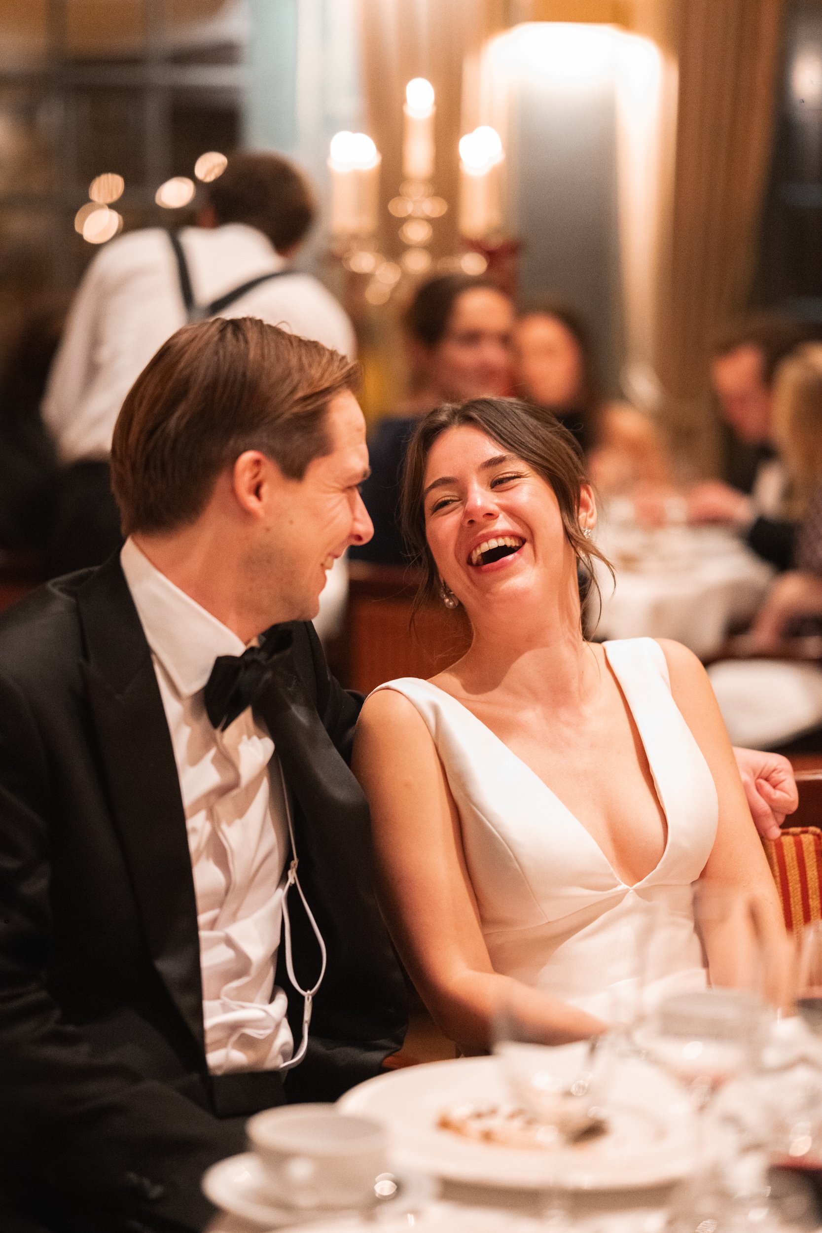 A smiling woman and man dressed in formal attire, wedding dress and tuxedo, sharing a joyful moment at a wedding reception.