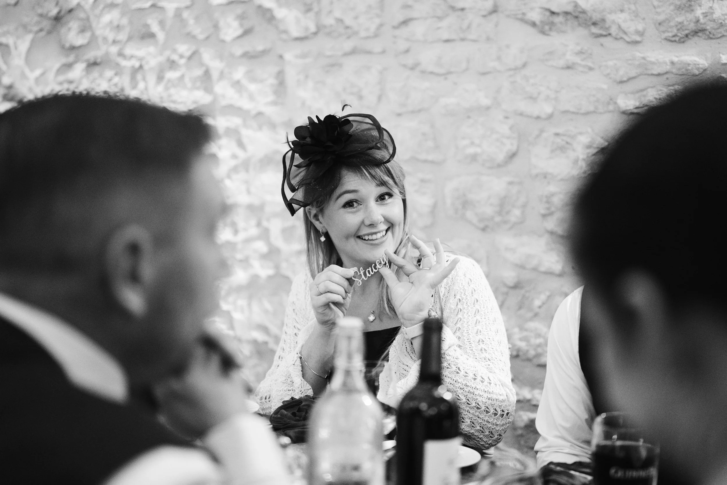 A woman wearing a fascinator hat smiling at a celebration, holding a decorative sign that says "Stacey," with other people and bottles on the table.