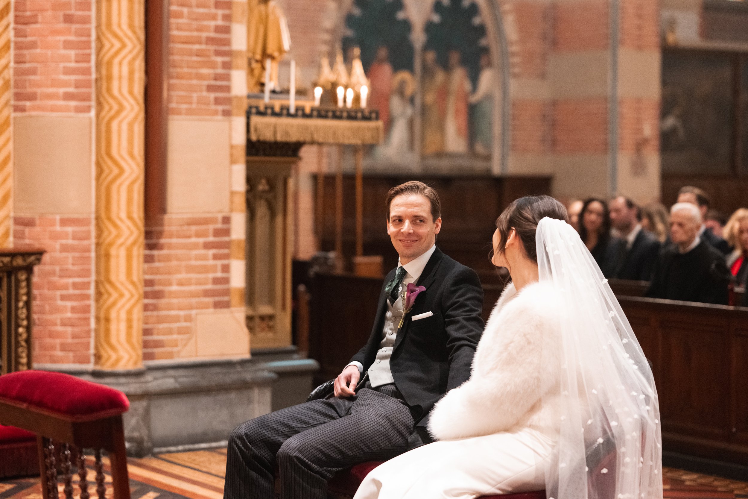 A bride and groom sitting in a church during their wedding ceremony. The groom is smiling and dressed in a black suit with a purple boutonniere. The bride, wearing a white veil and fur shawl, is sitting beside him.