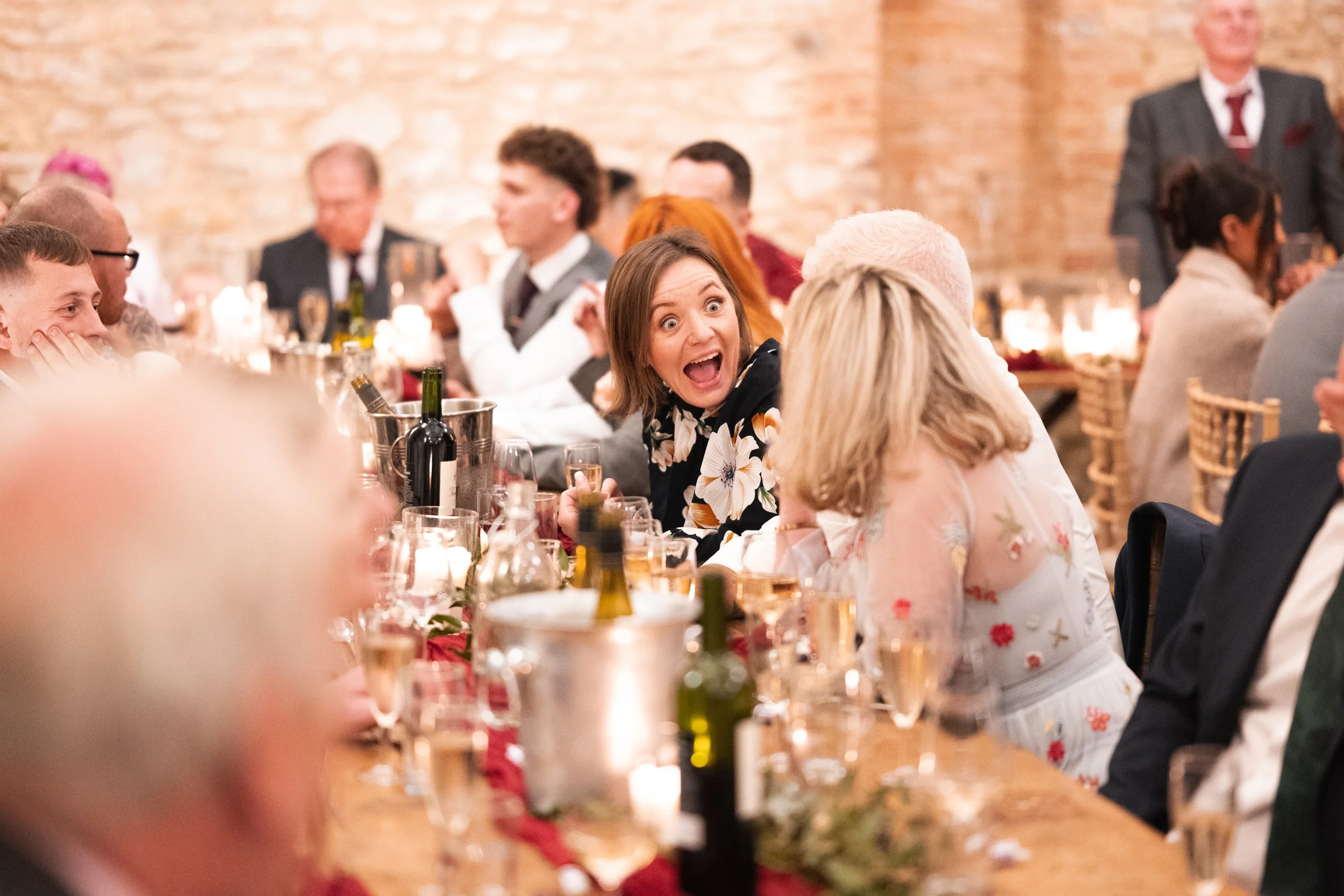 A woman with shoulder-length brown hair and wearing a floral dress, sitting at a dinner table, has an expressive surprised or excited look on her face, talking to the woman in front of her, with other guests seated at the table in the background.