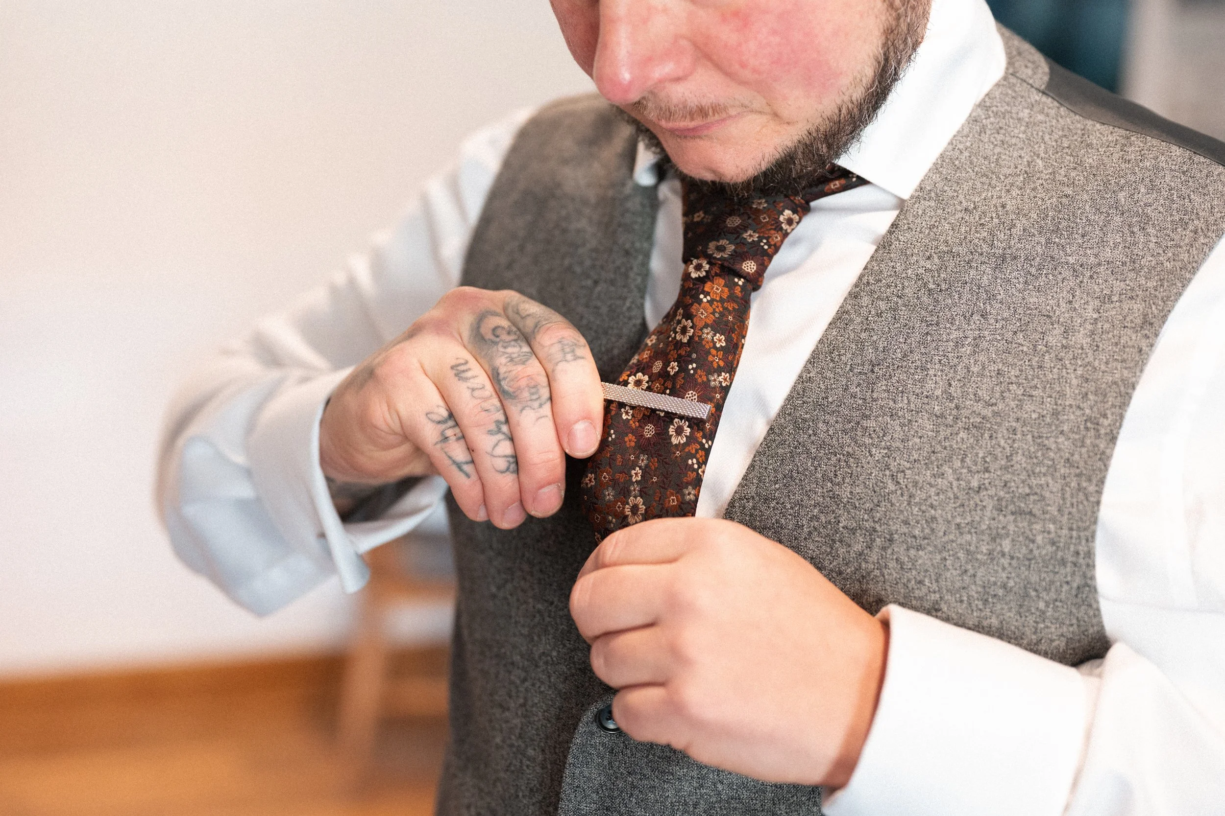 A man with tattoos on his hand adjusting a floral-patterned tie while wearing a white shirt and grey vest.