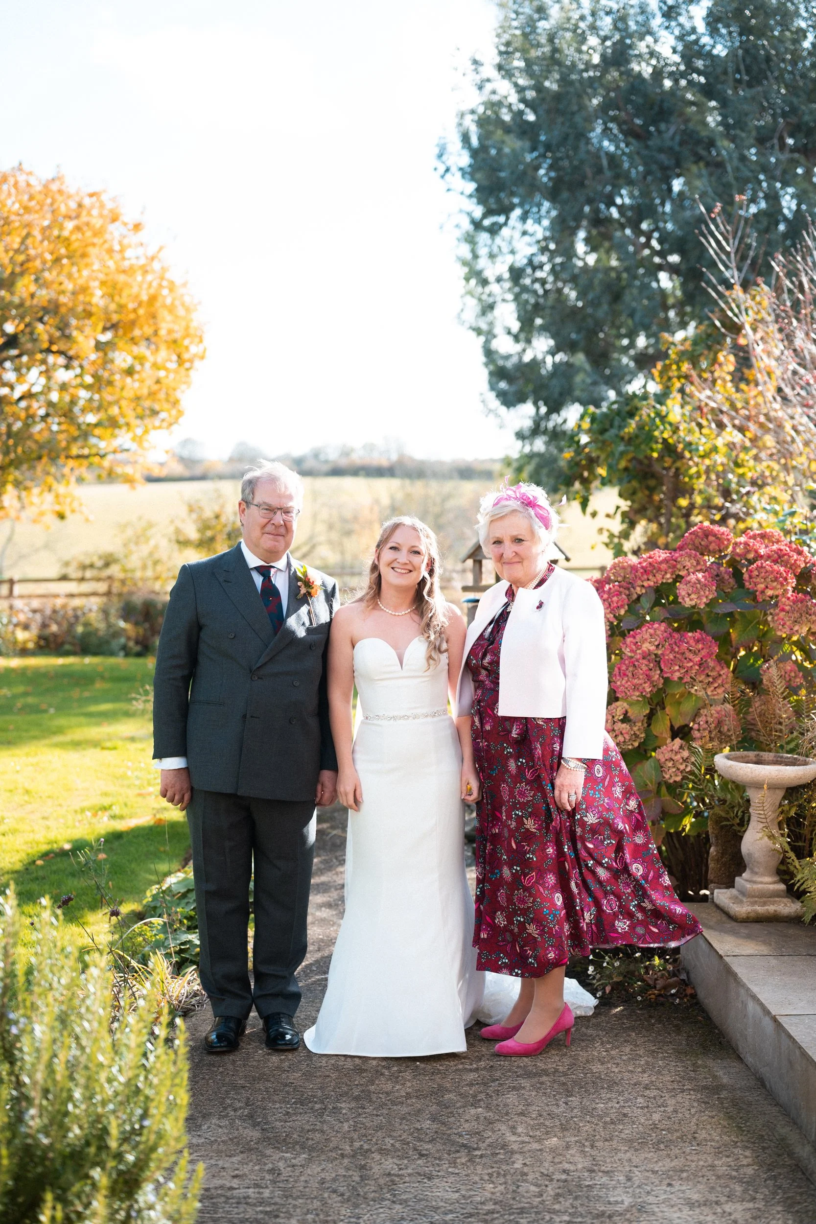 Three people standing outdoors in a garden on sunny day, a woman in a white wedding dress in the middle, flanked by a man in a dark suit and a woman in a floral dress and pink heels.