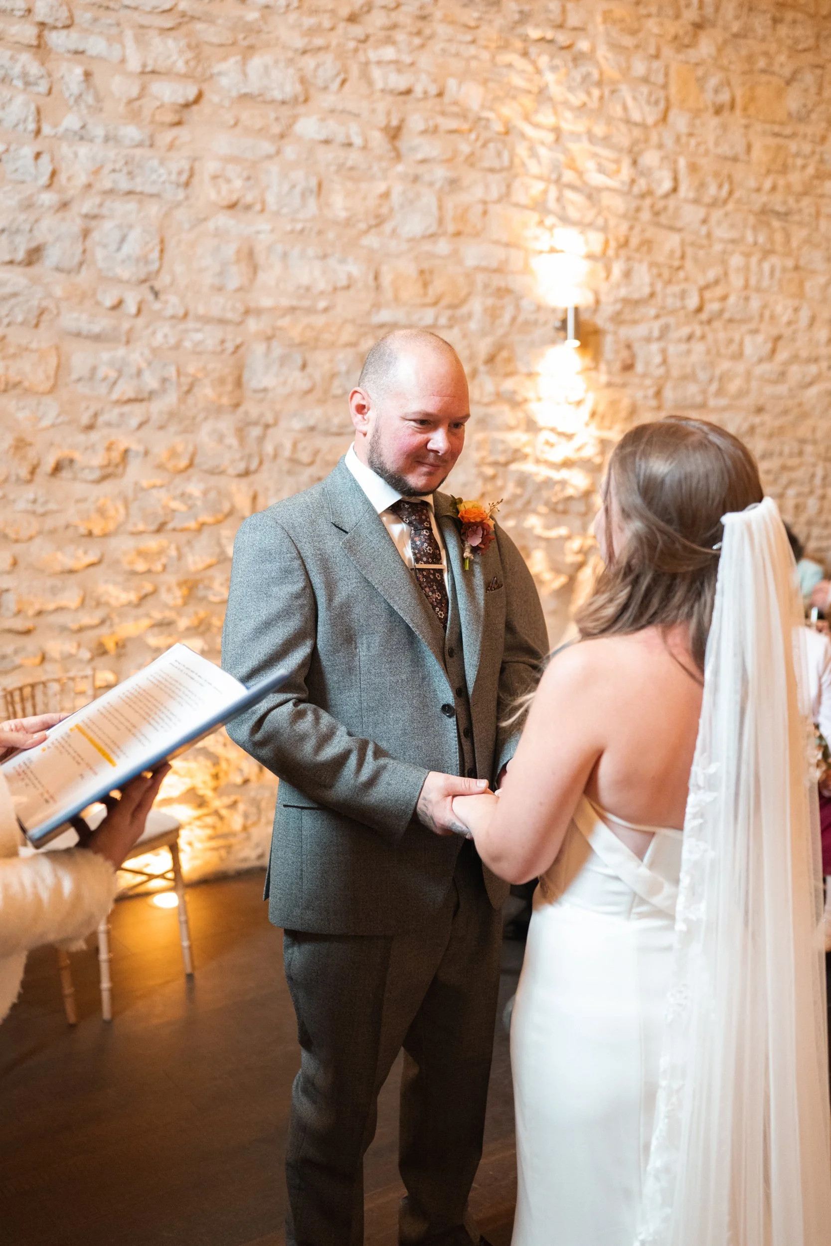 A groom and bride holding hands during a wedding ceremony in a rustic setting with stone walls.