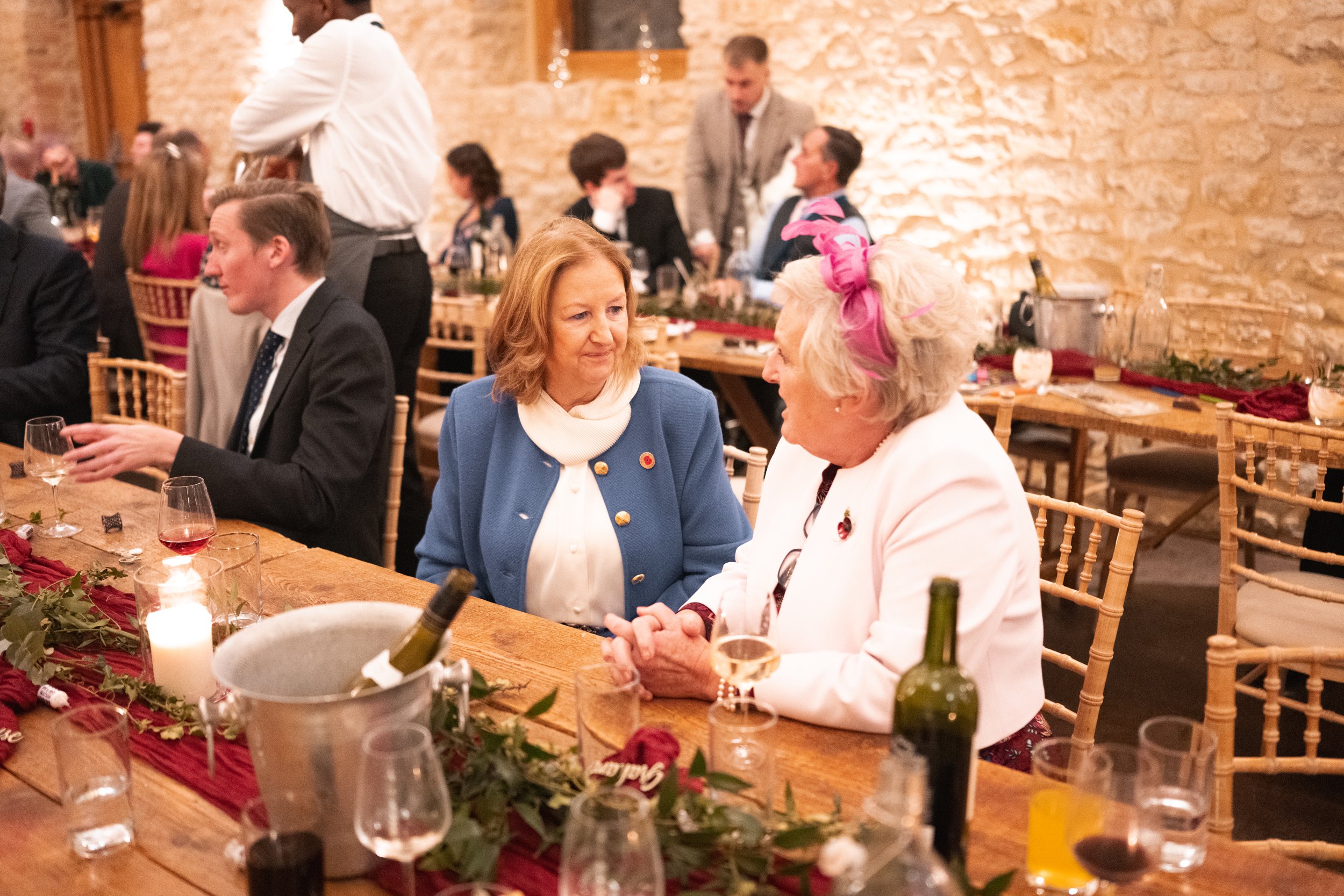 Two women having a conversation at a decorated dinner table in a warmly lit brick-walled venue, with other guests in the background.