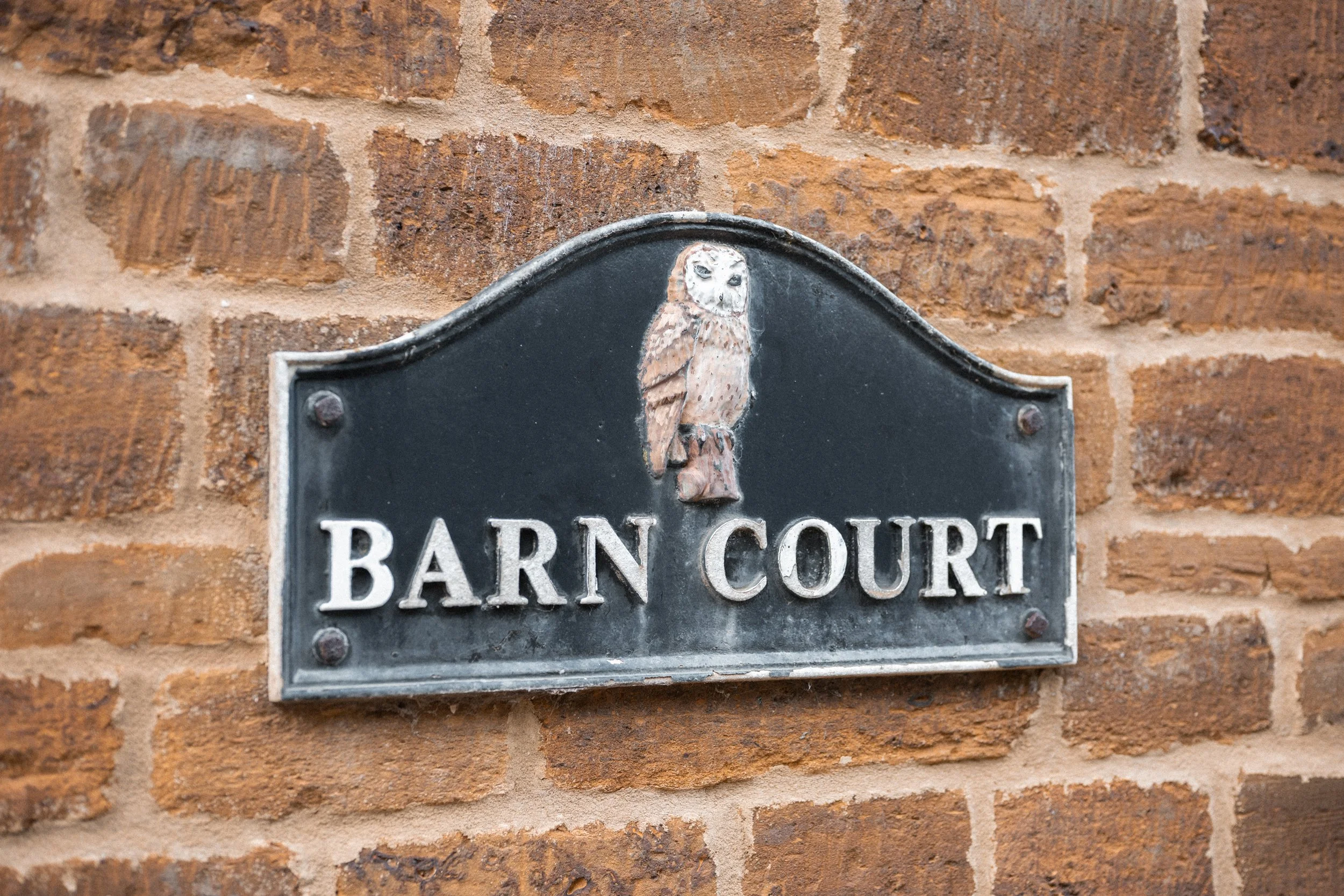 A black metal street sign with a painted owl at the top, mounted on a brick wall, reading "BARN COURT".