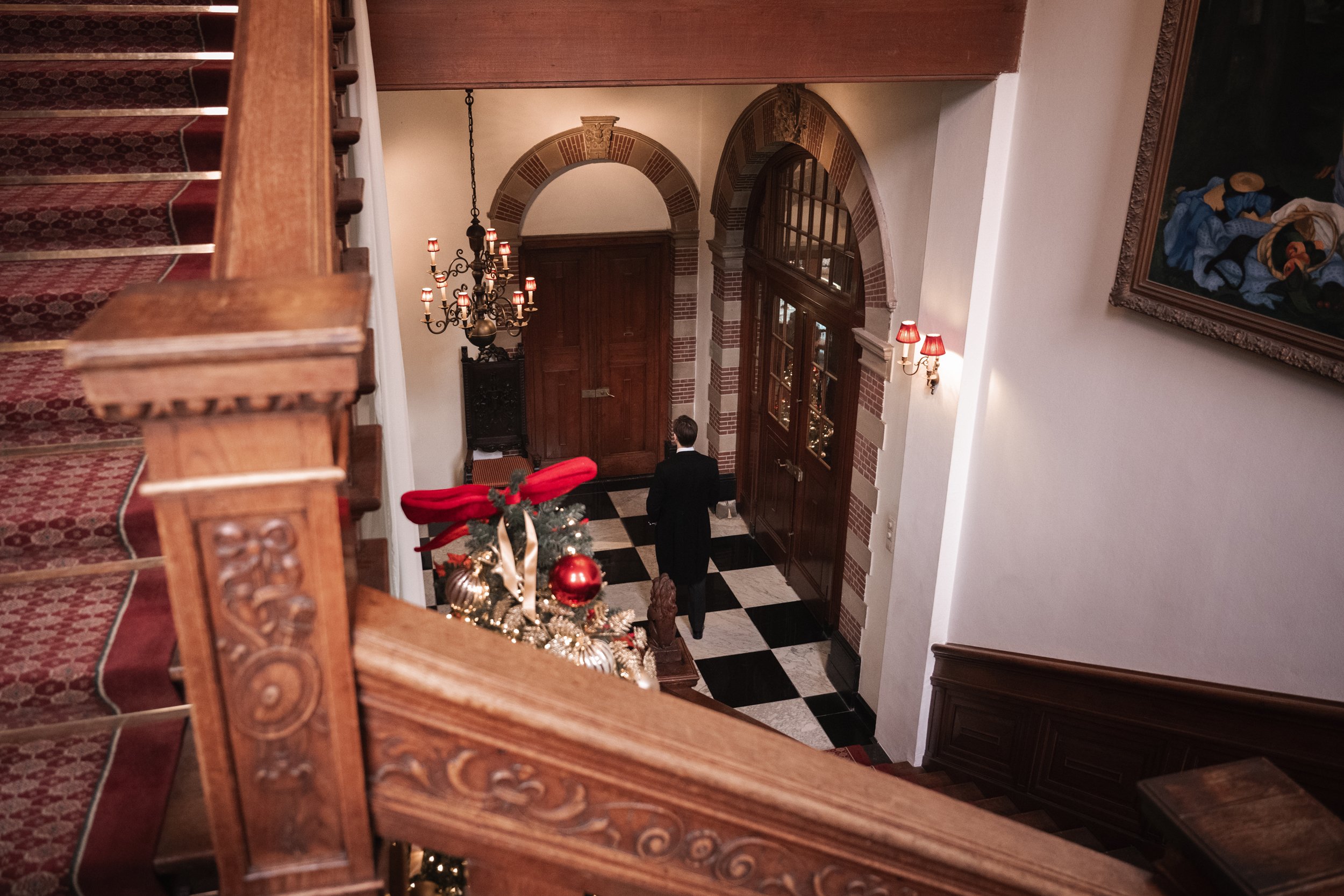 View of a hotel lobby or grand home entrance hall decorated for Christmas, with a stained-glass door, a chandelier, wall sconces, a decorated Christmas tree, and a man in a tuxedo walking toward the doorway.
