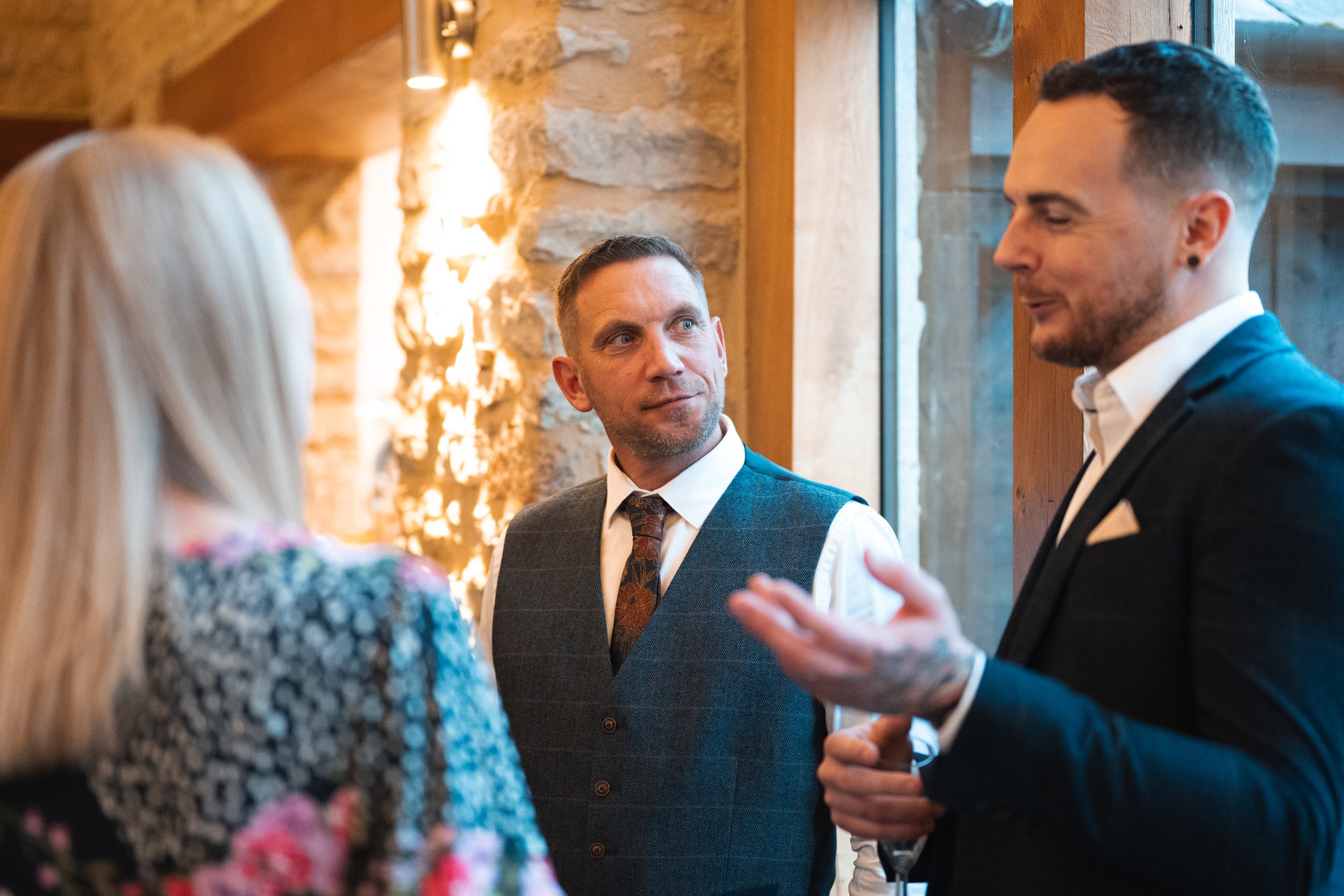 Three people engaged in conversation at an indoor social gathering, with a rustic stone wall and large window in the background.