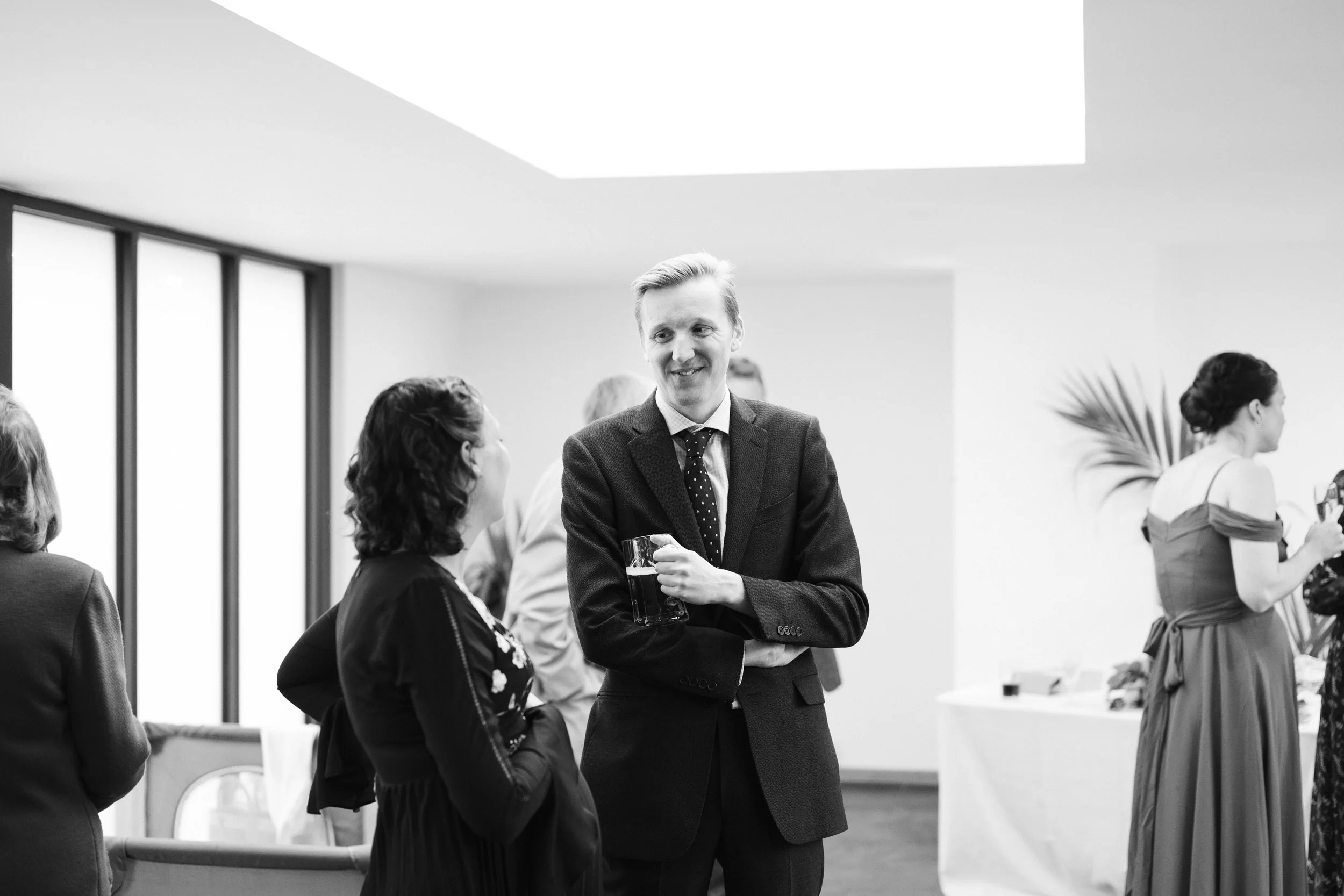 Black and white photo of a group of people at a social gathering. One man in a suit is smiling and holding a glass, talking to a woman with dark curly hair. Other women are engaged in conversation in the background.