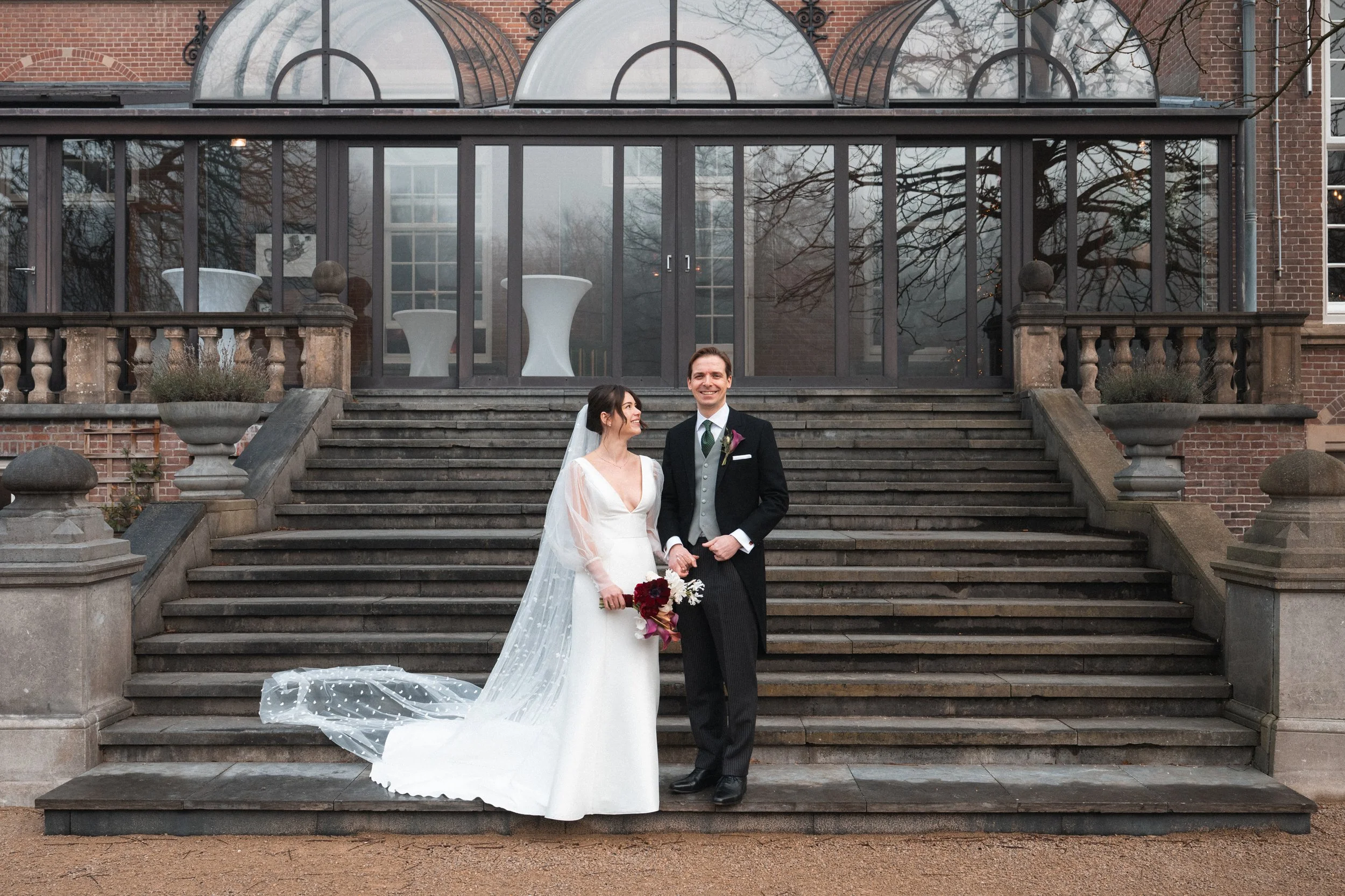 A bride and groom standing on outdoor stairs in front of a glass-encased building, holding hands and smiling, during their wedding.
