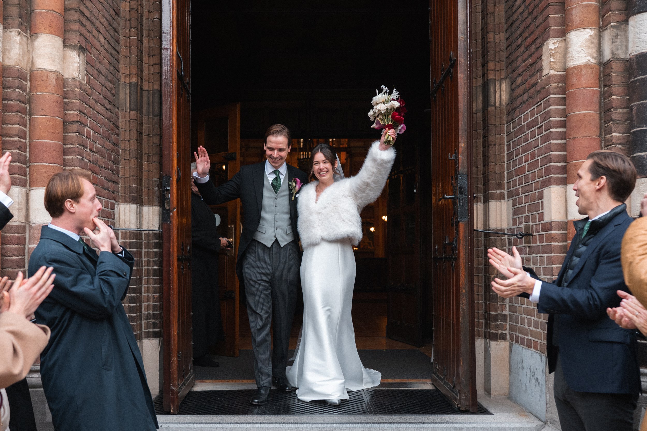 A newly married couple exiting a church; the bride holds a bouquet and raises her arm in celebration, while the groom smiles and waves, surrounded by applauding guests.