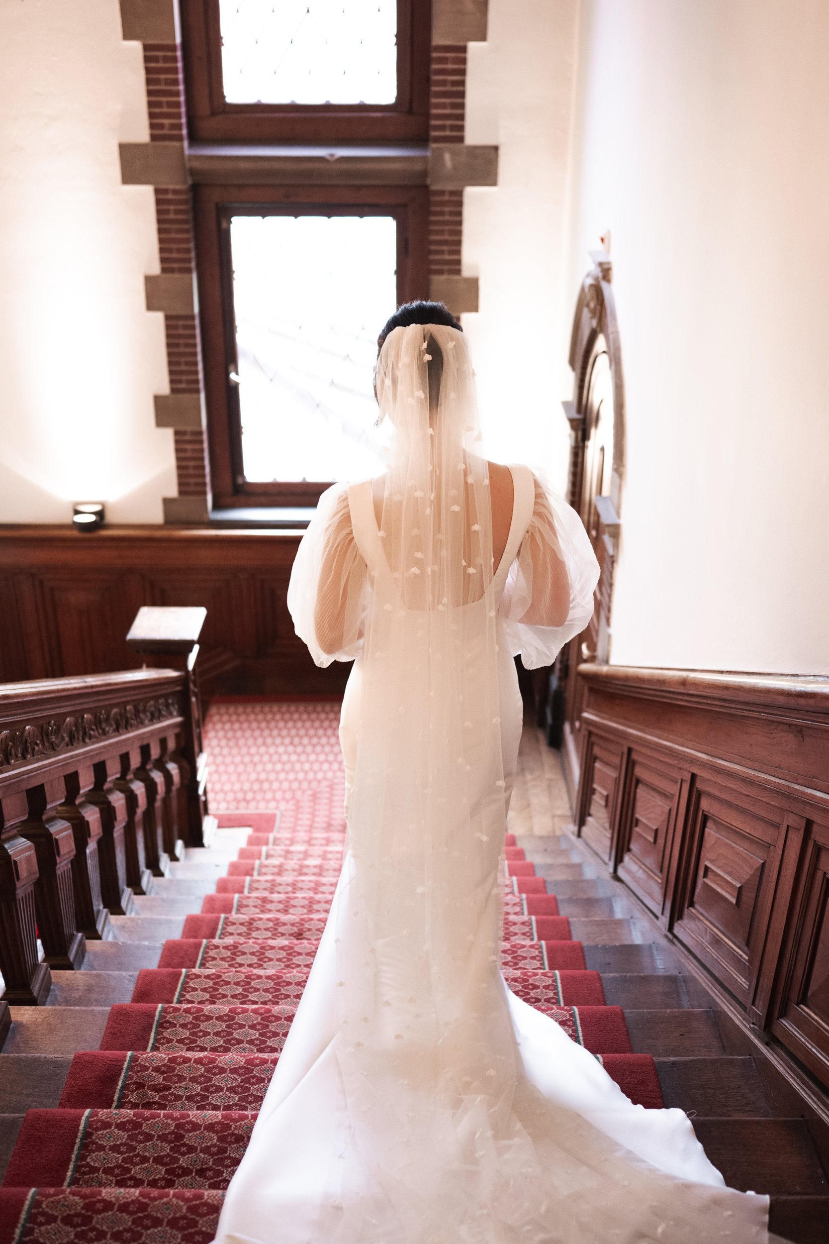 A bride in a white wedding dress with puffed sleeves and a long veil stands on a staircase inside a building with wood-paneled walls and large windows.