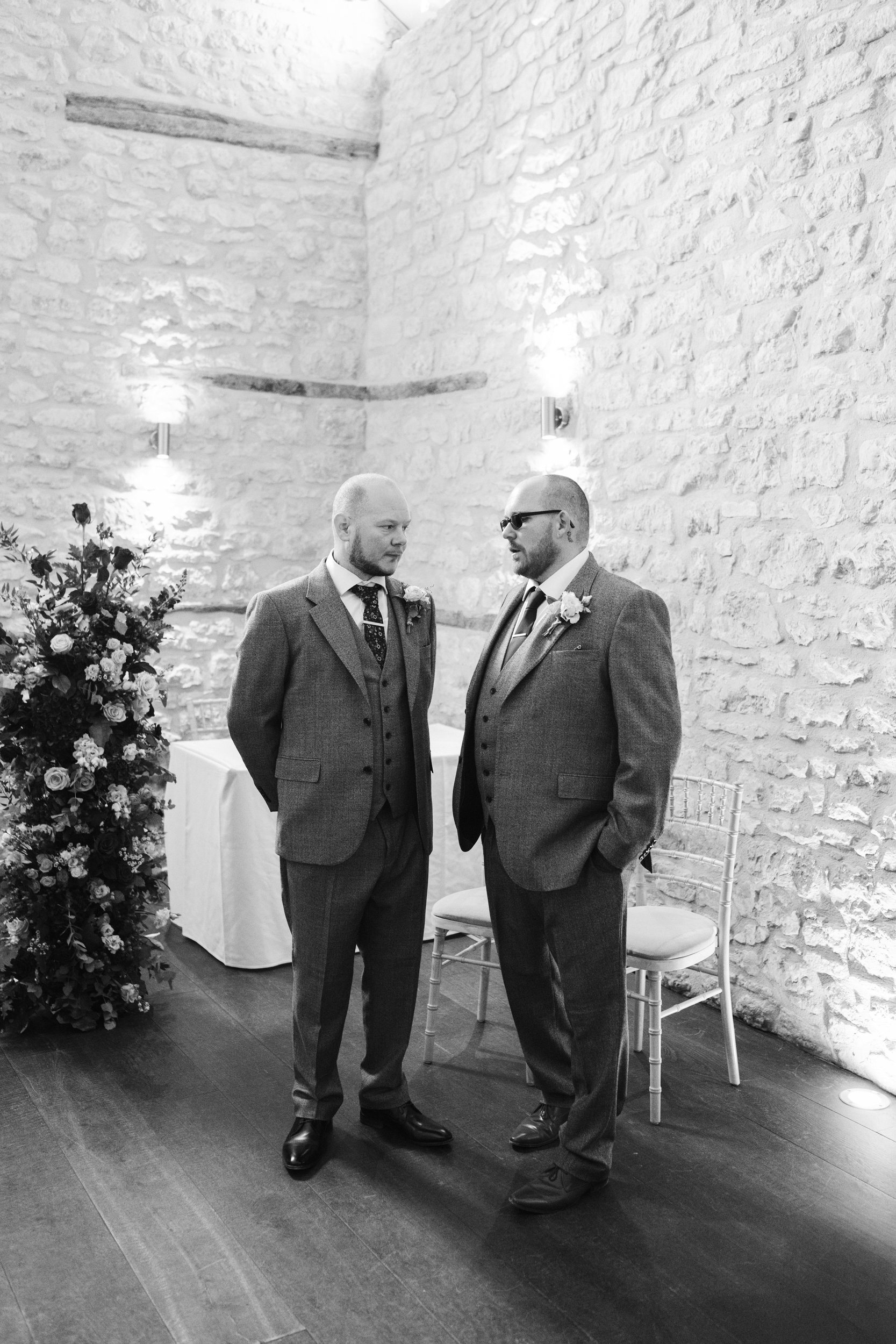 Two men in suits standing and talking indoors near a wall with ambient lighting. One man is wearing sunglasses, the other is not. There are chairs, a floral arrangement, and a table in the background.
