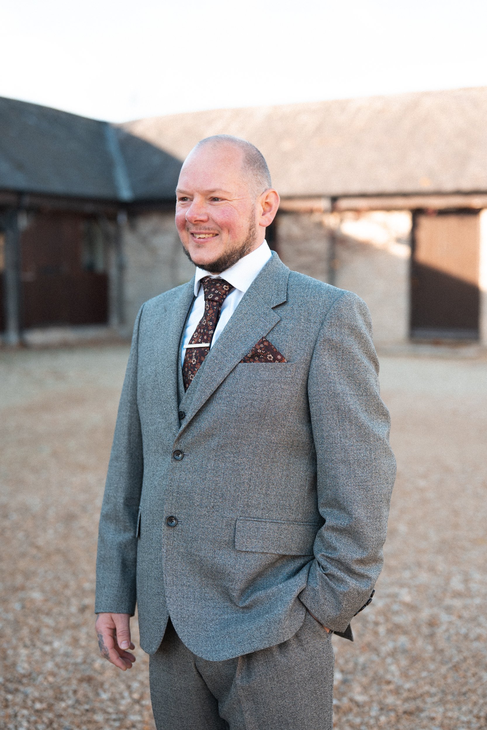 A man in a gray suit with a patterned tie and pocket square, standing outdoors on a gravel area, smiling, with rustic buildings in the background.