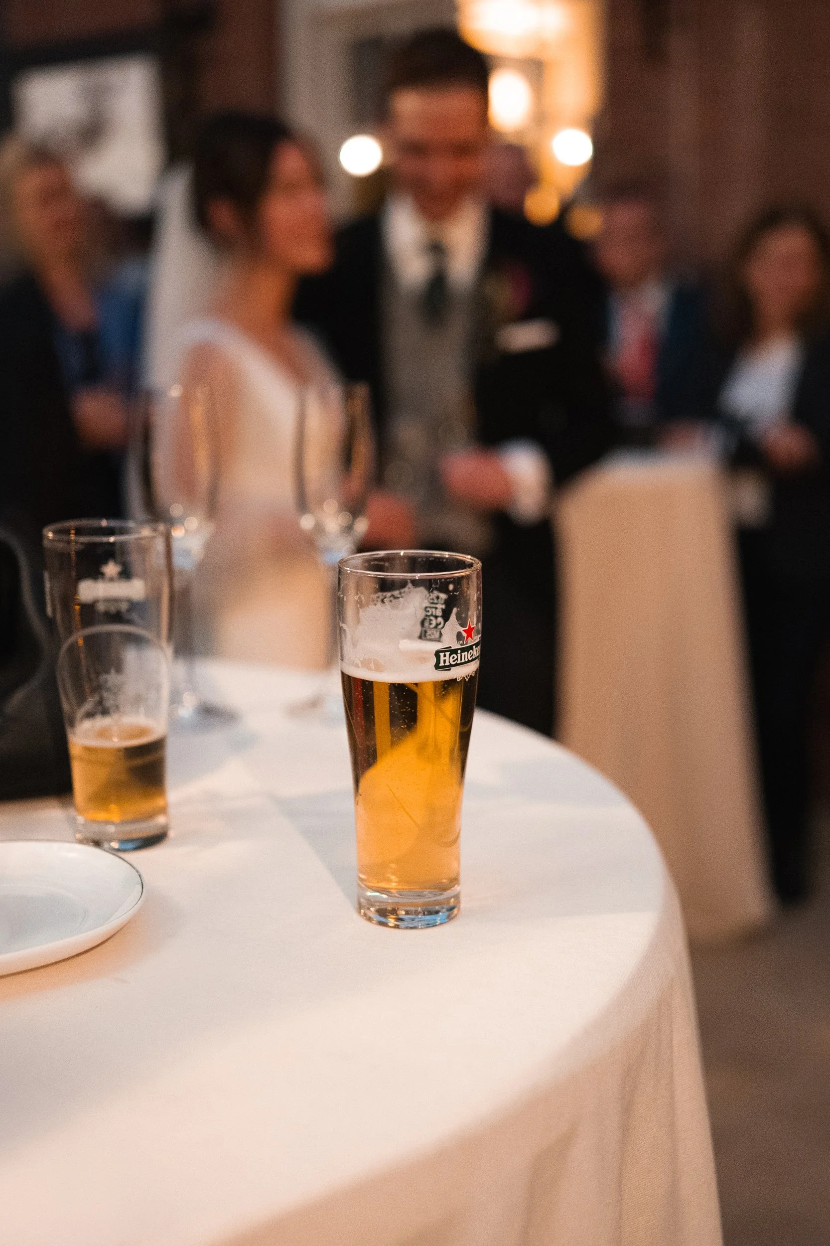 A glass of beer on a round table at a wedding reception, with wedding guests blurred in the background.