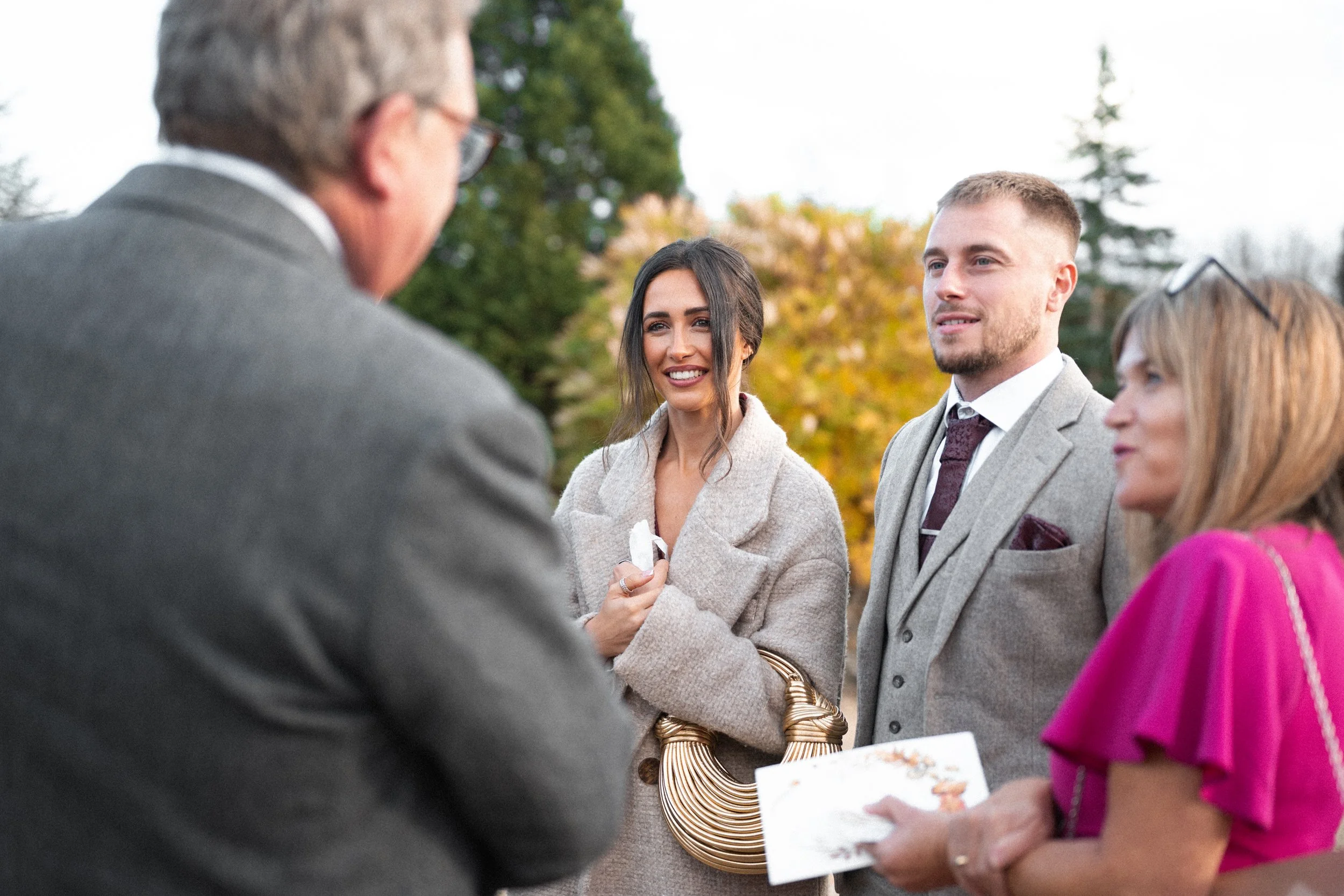 A group of four people standing outdoors, engaged in conversation. One man wearing glasses and a grey suit is talking to a woman in a beige coat, holding a small white object. Next to her is a young man in a grey suit and white shirt. An older woman 