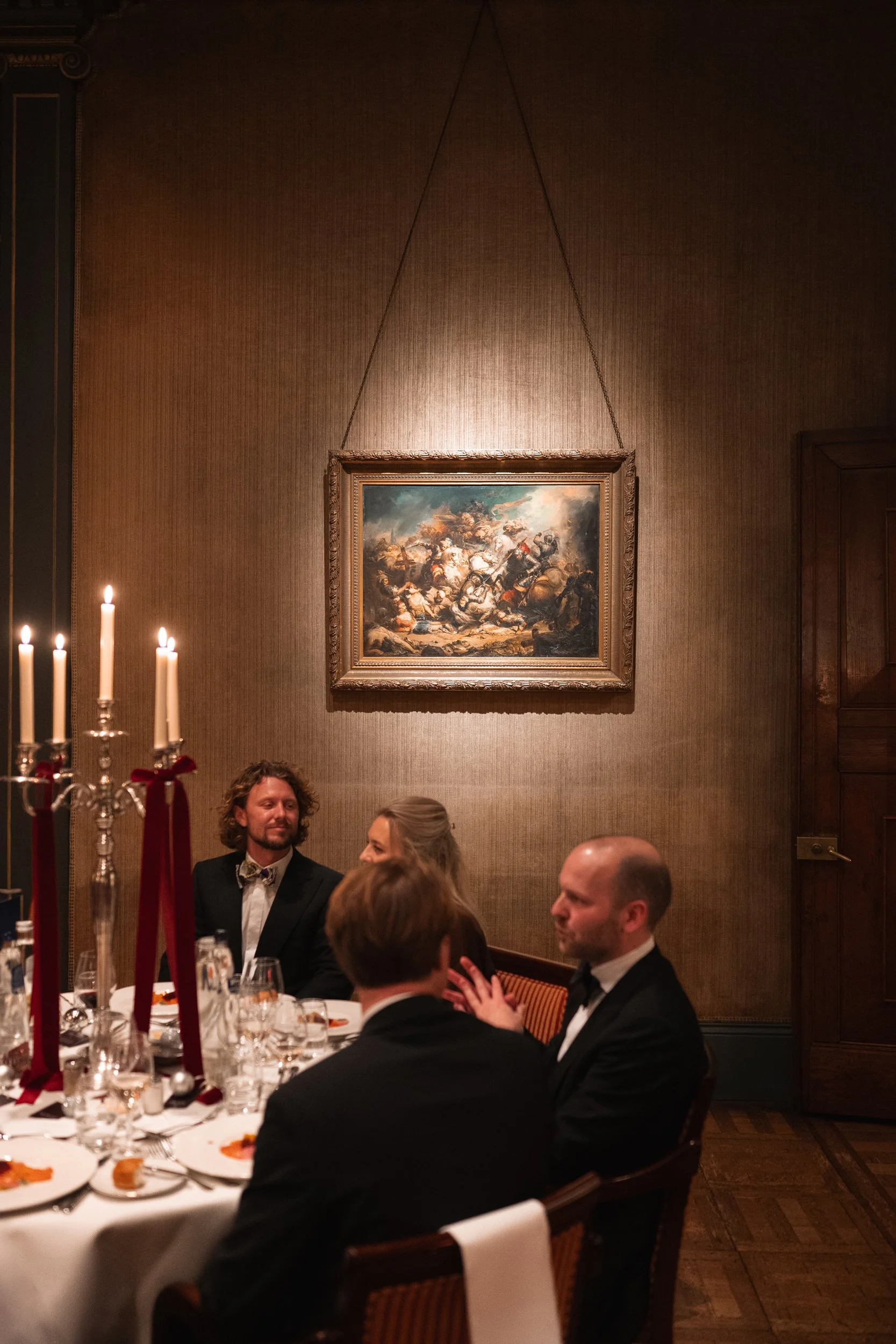 People dressed in formal attire sitting at a dinner table in a dimly lit room with a large painting on the wall.