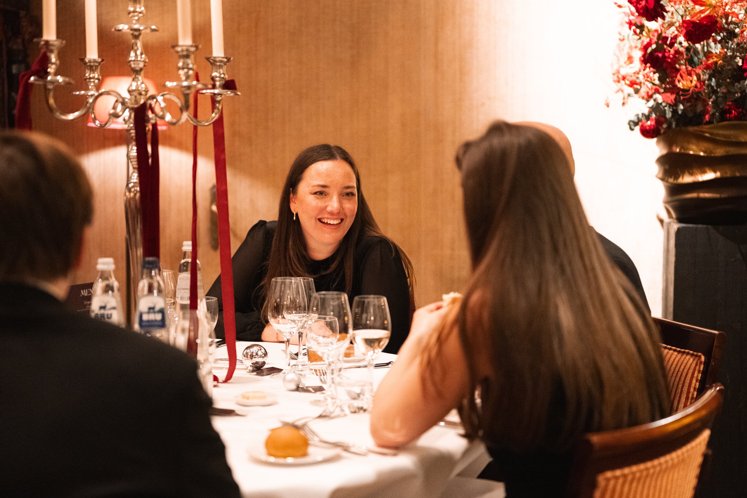 Group of four people sitting around a dining table, enjoying conversation at a festive event, with ornate candle holder and floral arrangements in the background.