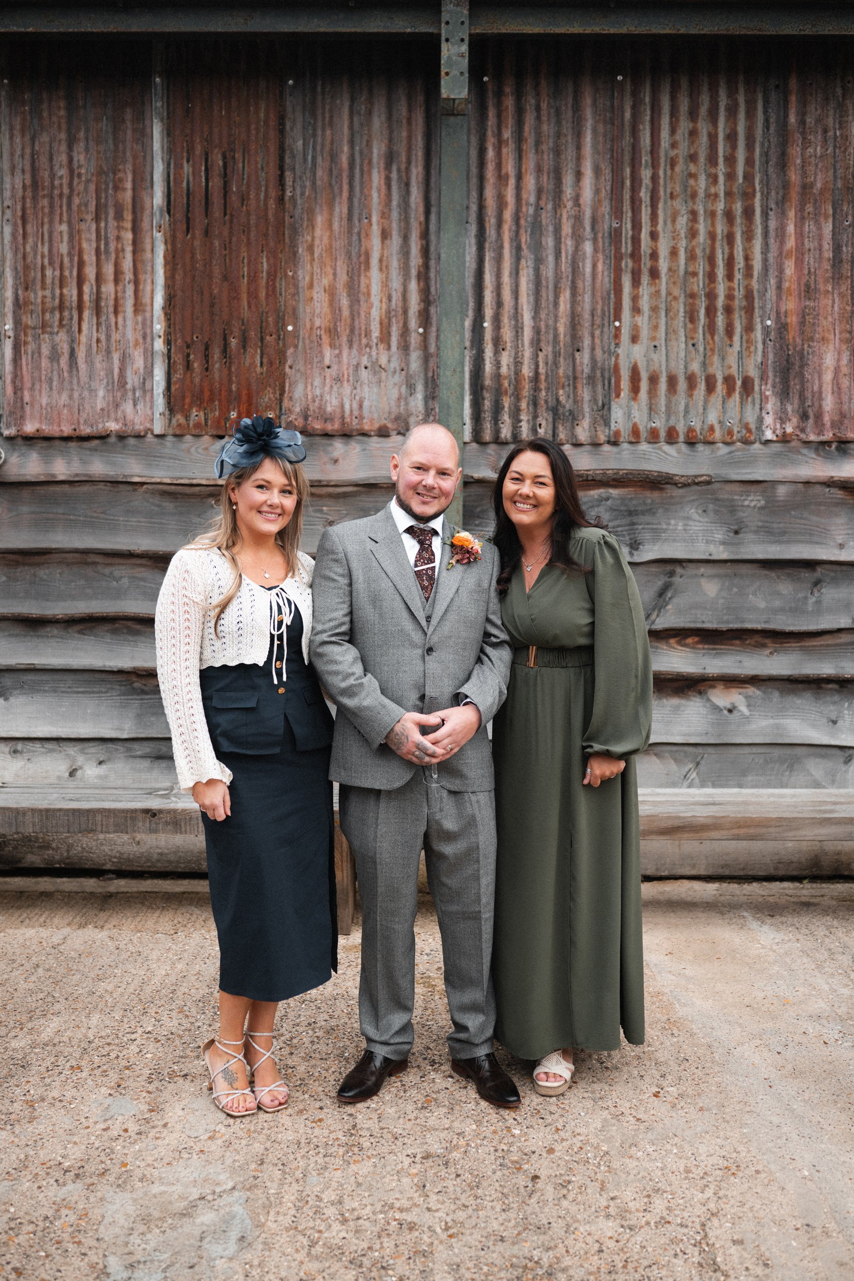 Three adults standing together in front of a rustic wooden barn, dressed in formal attire for a special occasion, smiling at the camera.