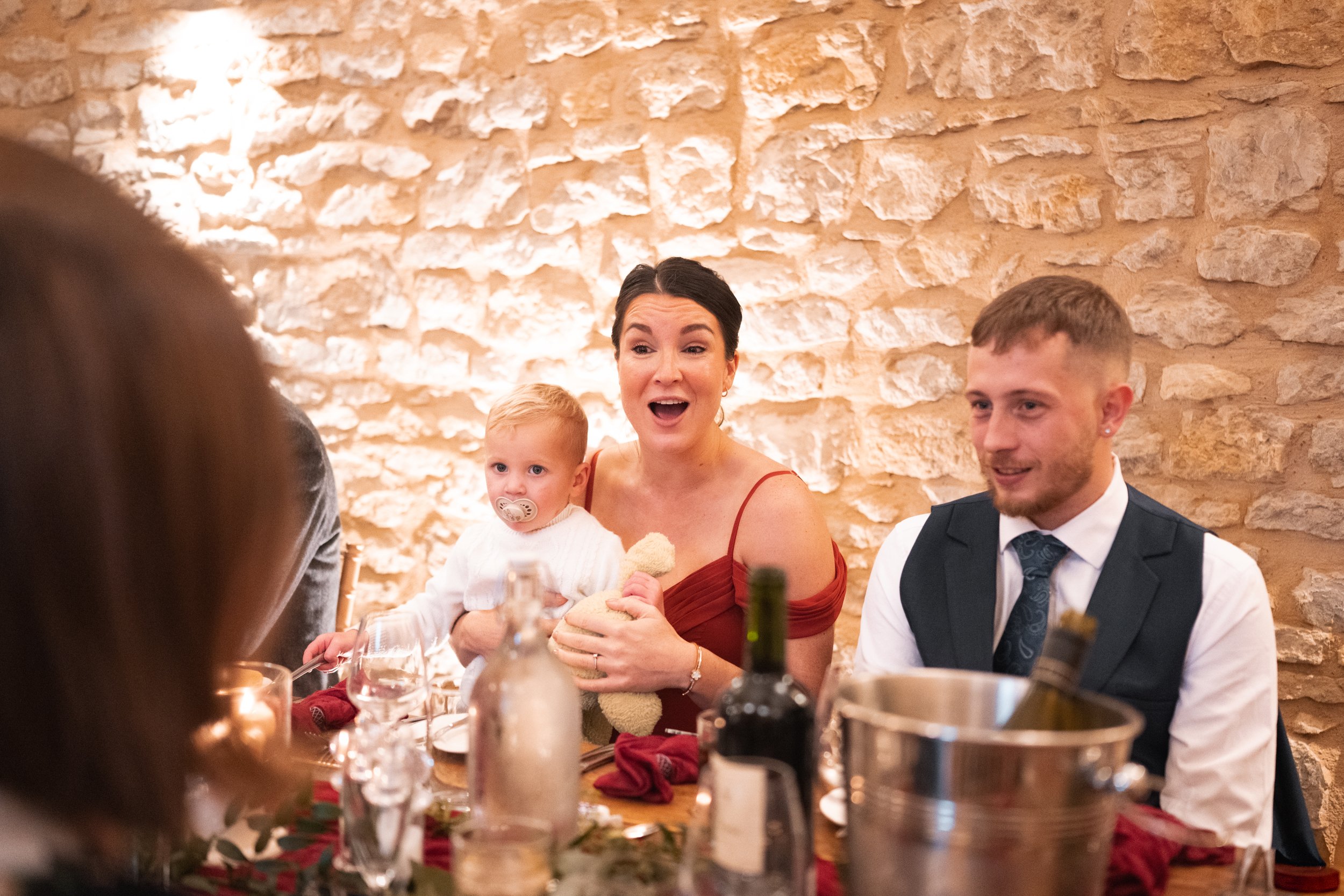 A woman in a red dress, a man in a suit, and a young child with a pacifier sit at a table during a celebration, with a brick wall in the background.