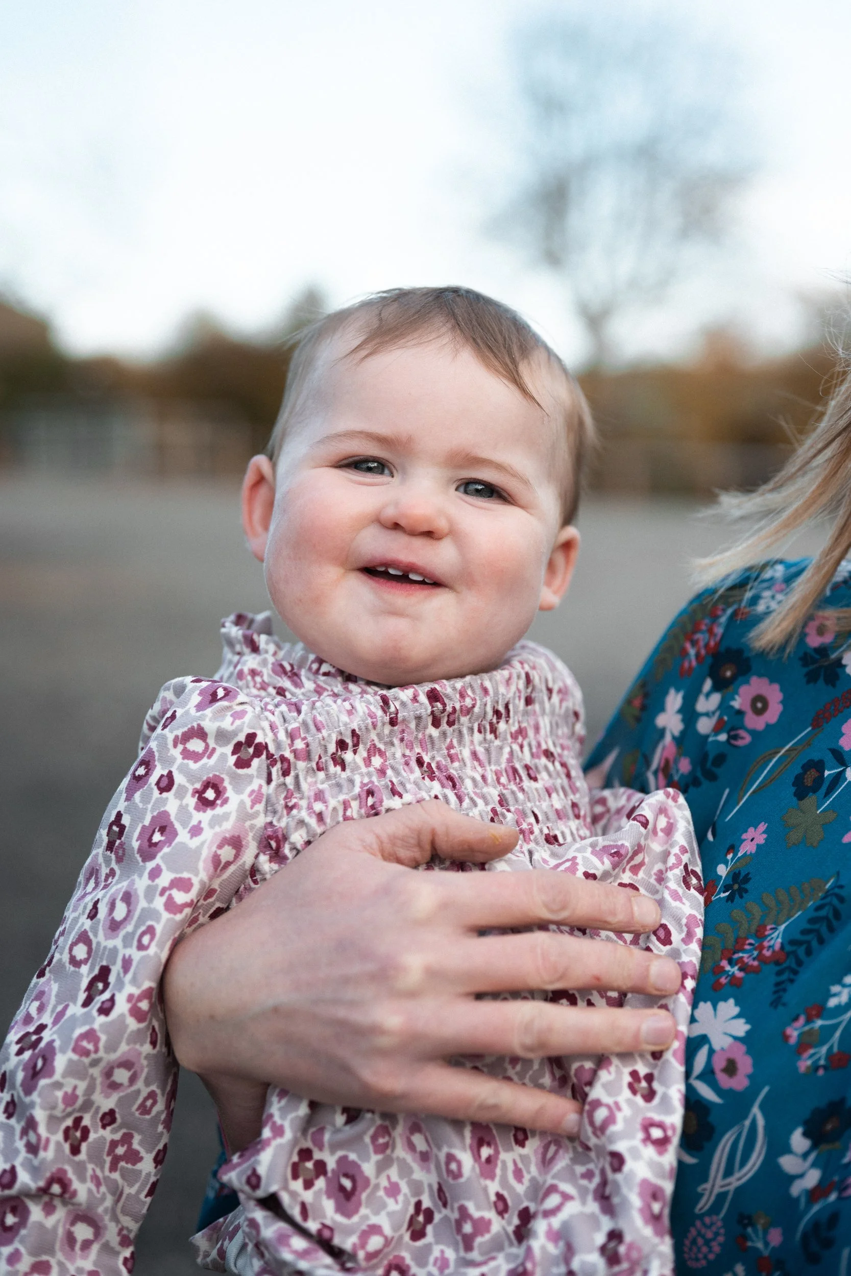 A smiling toddler girl with short brown hair, wearing a pink and white floral dress, being held by an adult in a blue floral shirt outdoors during daytime.