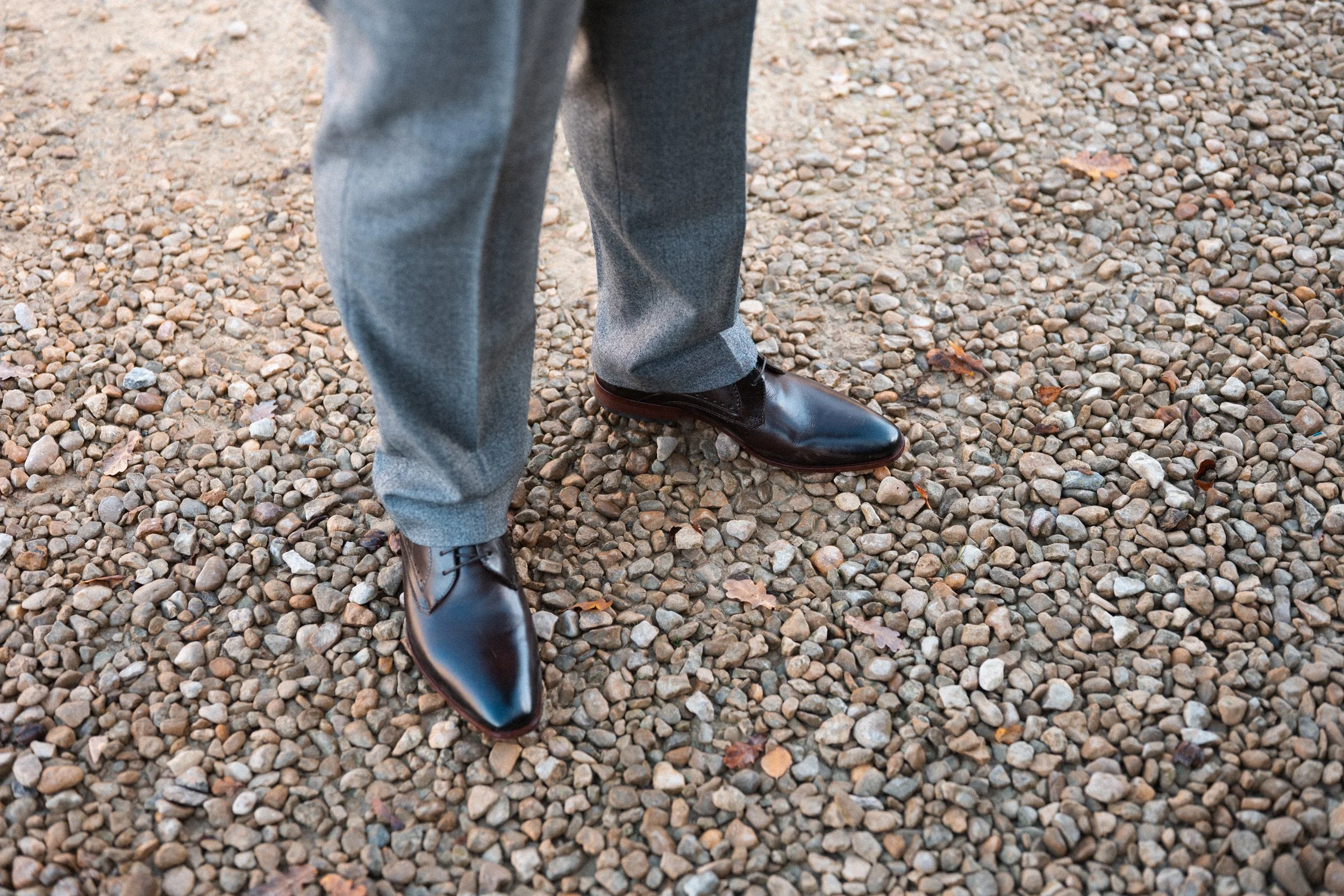 Person wearing gray trousers and black polished dress shoes standing on gravel surface.